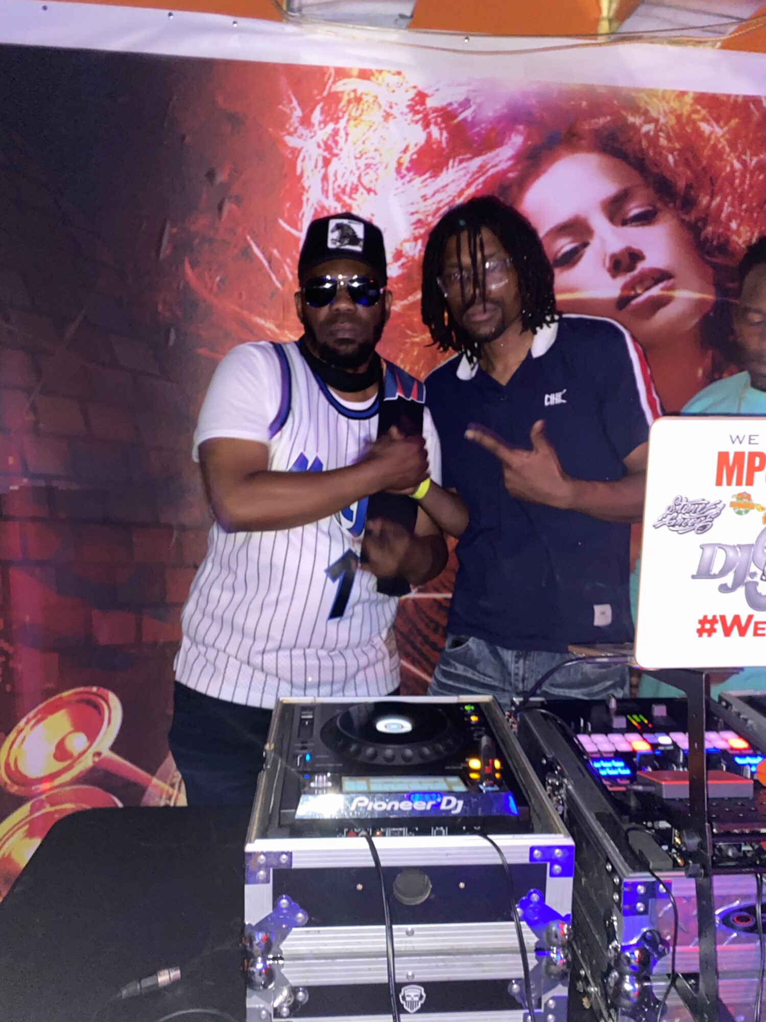 Two people standing behind DJ equipment, one holding a microphone, with a colorful background and a poster of a woman's face.