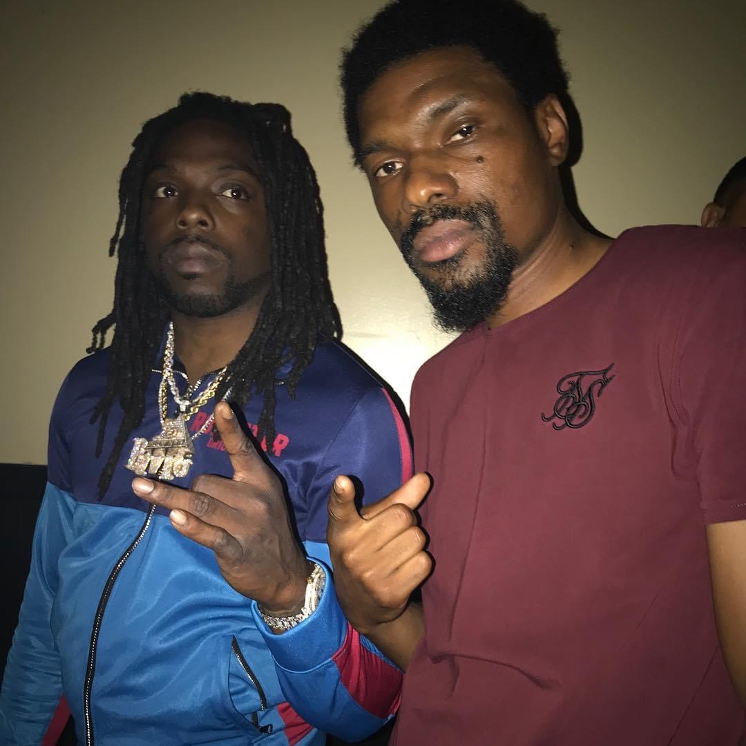 Two men pose together, one wearing a blue jacket with jewelry, the other in a maroon shirt, both making hand gestures.
