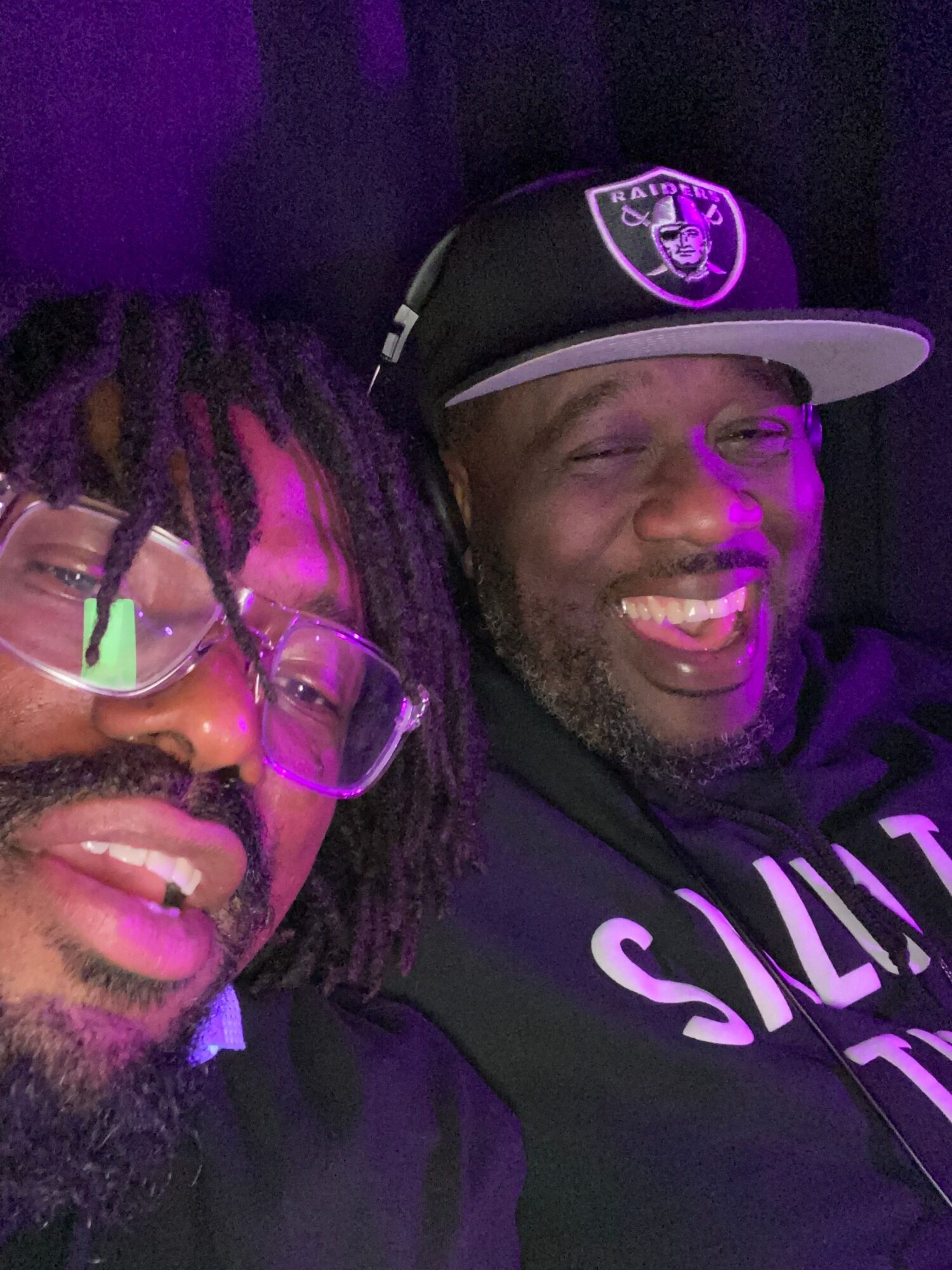 Two smiling men lying down, one wearing glasses and a cap, the other with dreadlocks and glasses, illuminated by purple light.