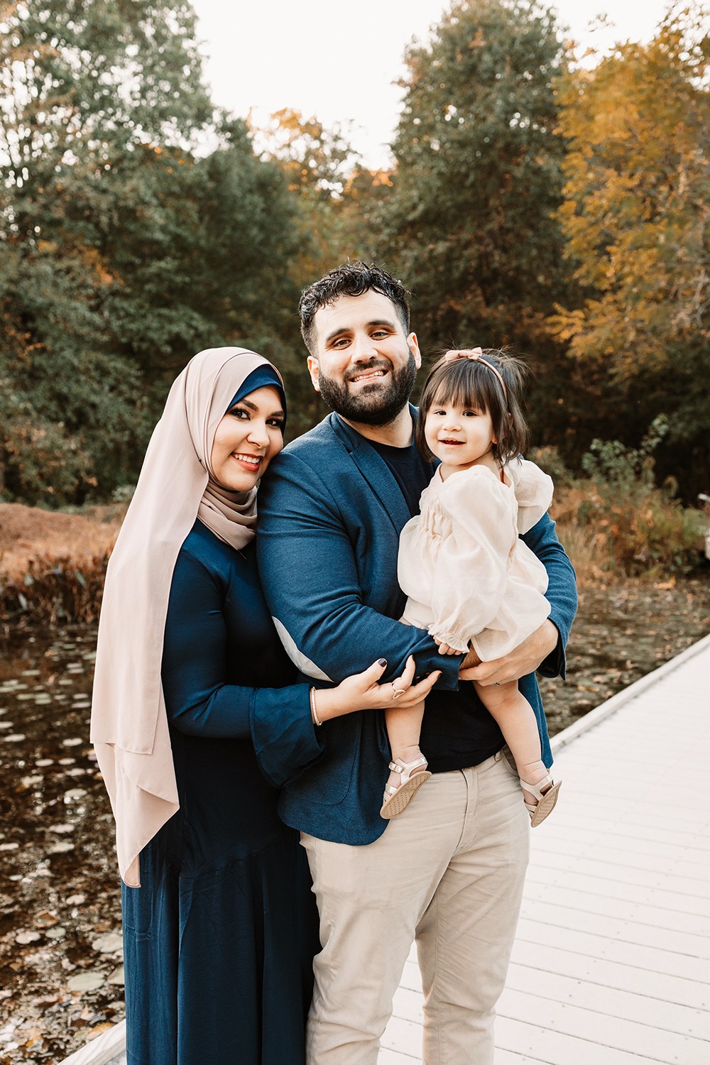 Family of three outdoors on a wooden path, smiling, with trees in autumn background.