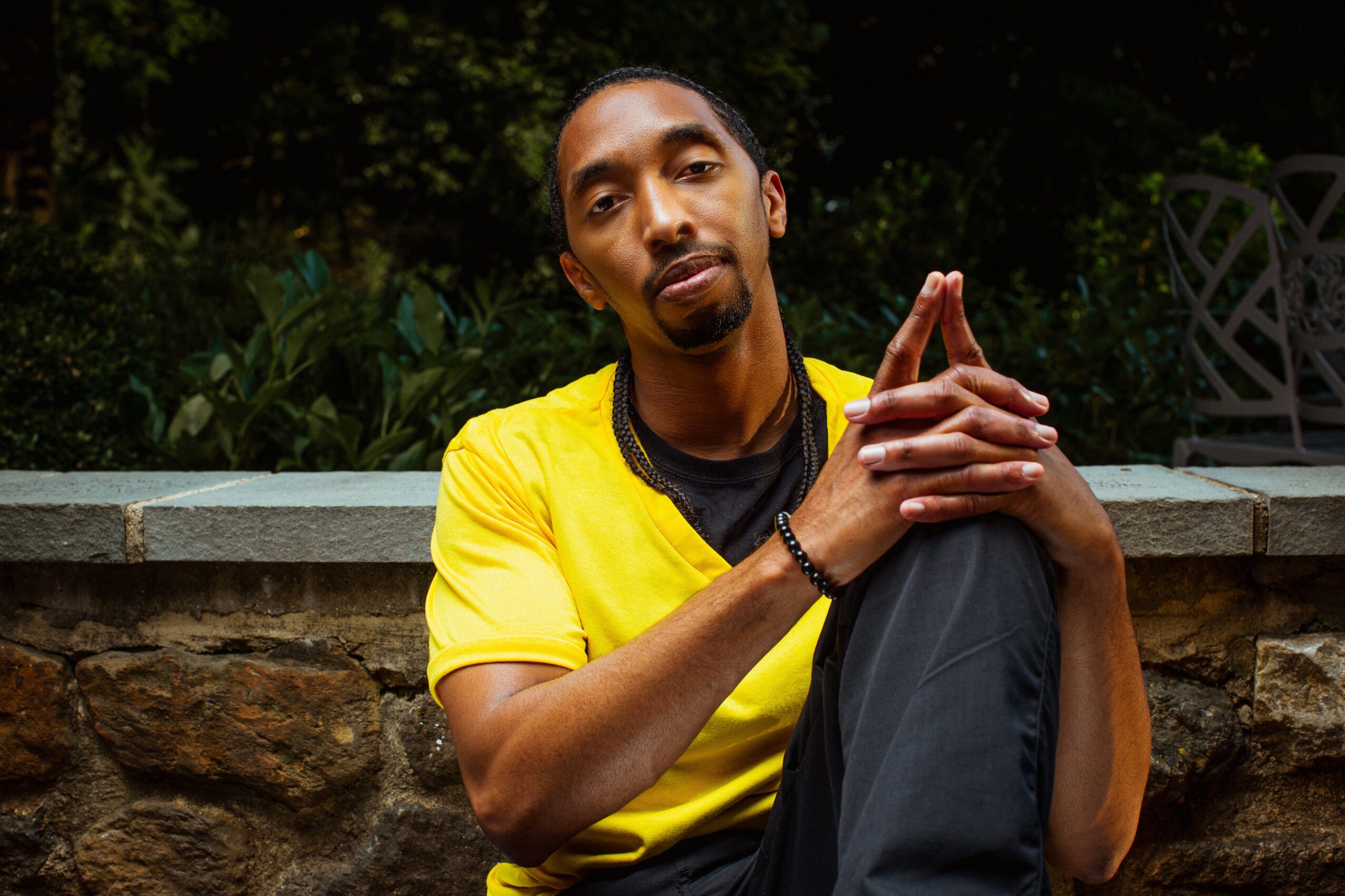 Man with long hair and a goatee wearing a yellow shirt, sitting outdoors with hands clasped, looking at the camera.