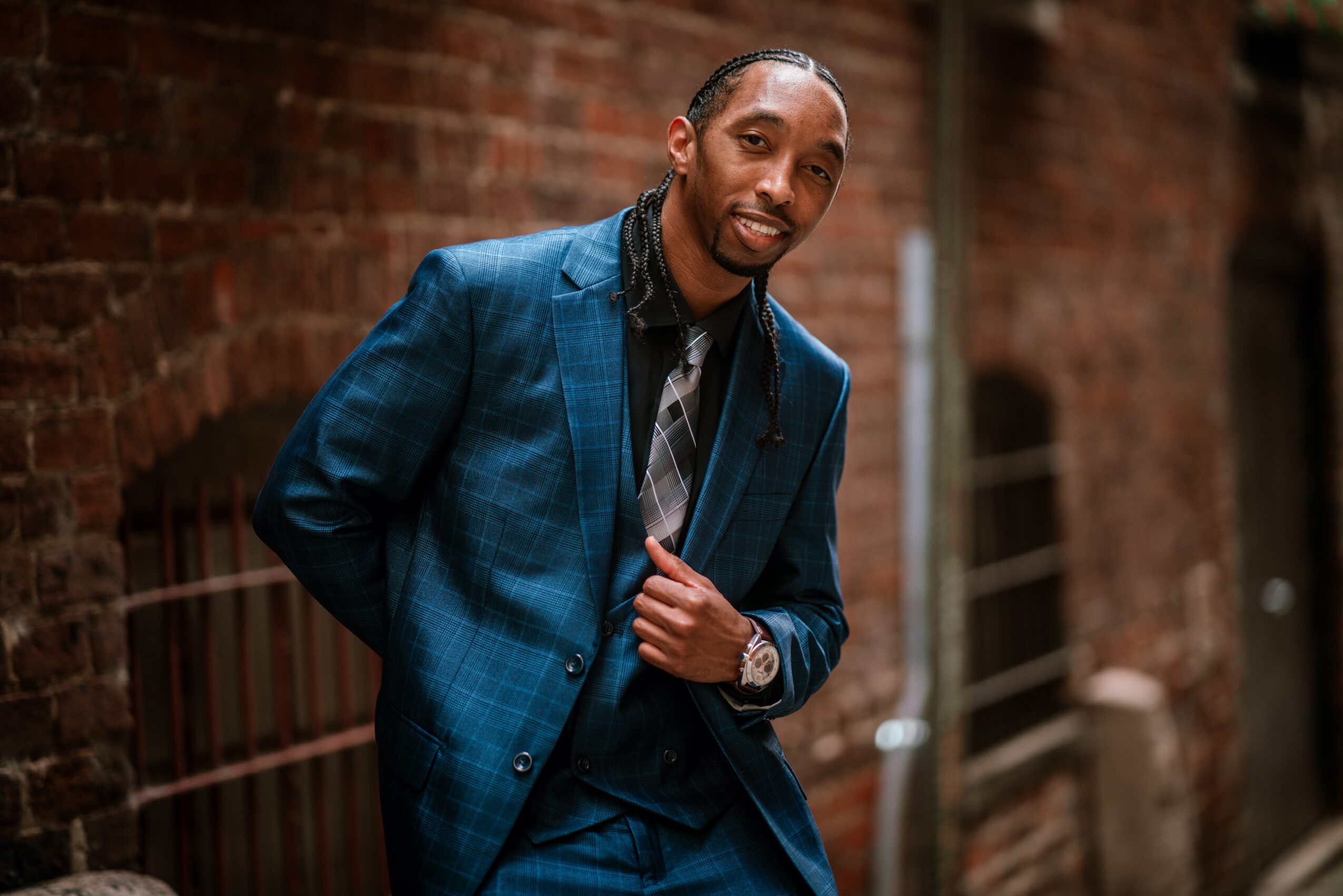 Man in a blue suit standing outdoors against a brick wall, smiling, with hands adjusting his jacket.