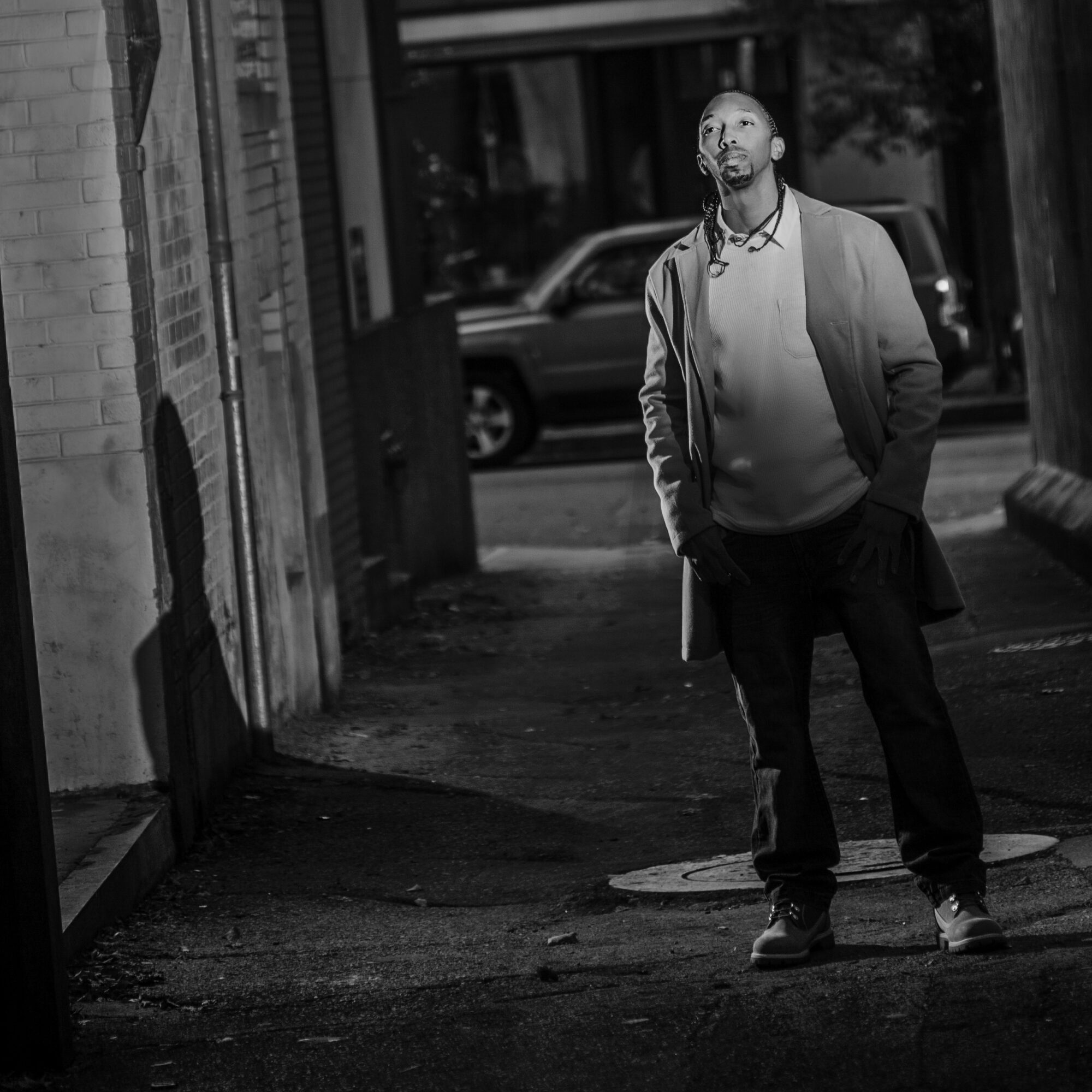 Man standing on sidewalk in urban area, looking upward, with parked car and building in background.