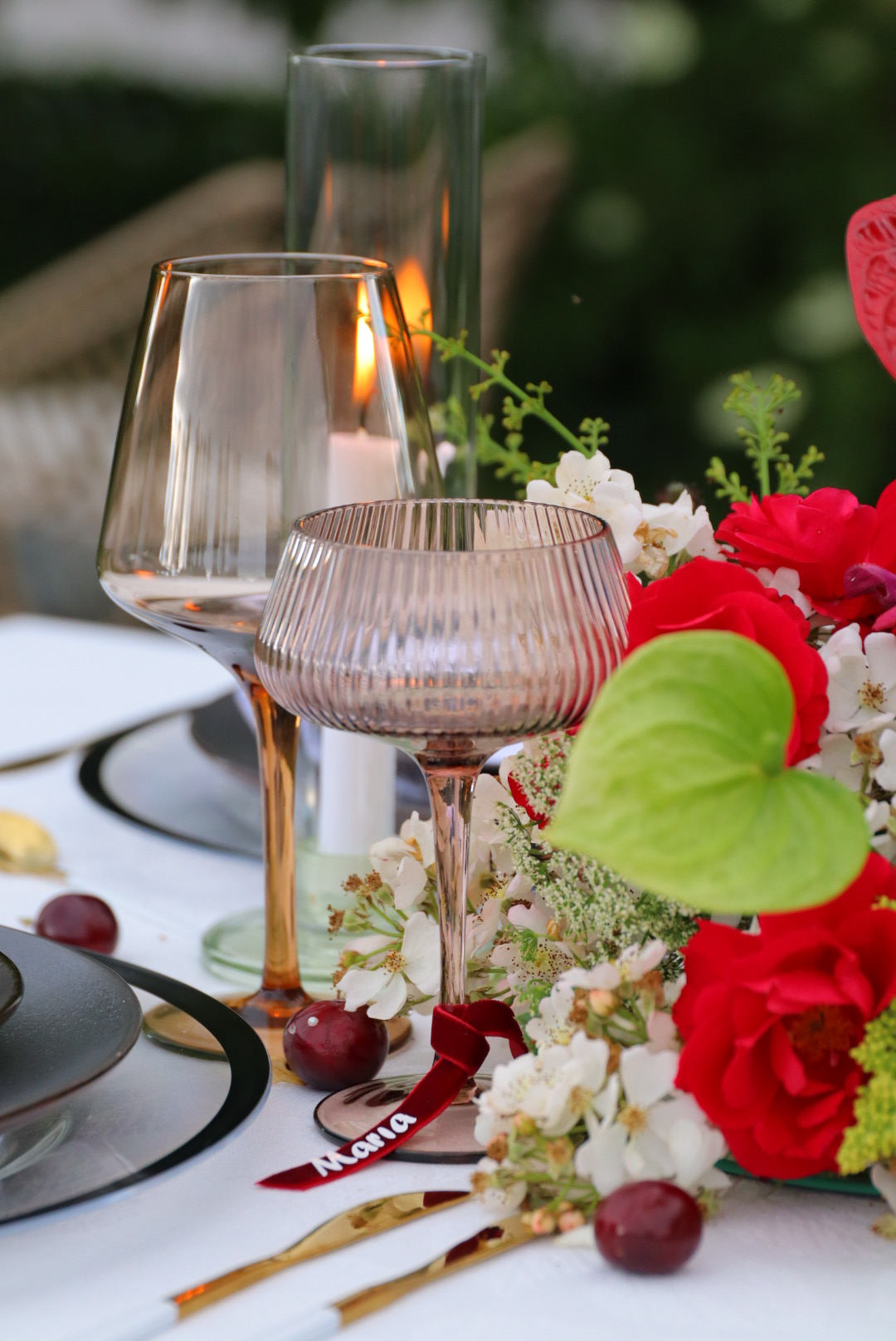 Glass pitcher pouring liquid into a glass, surrounded by red and white flowers, with greenery in the background.