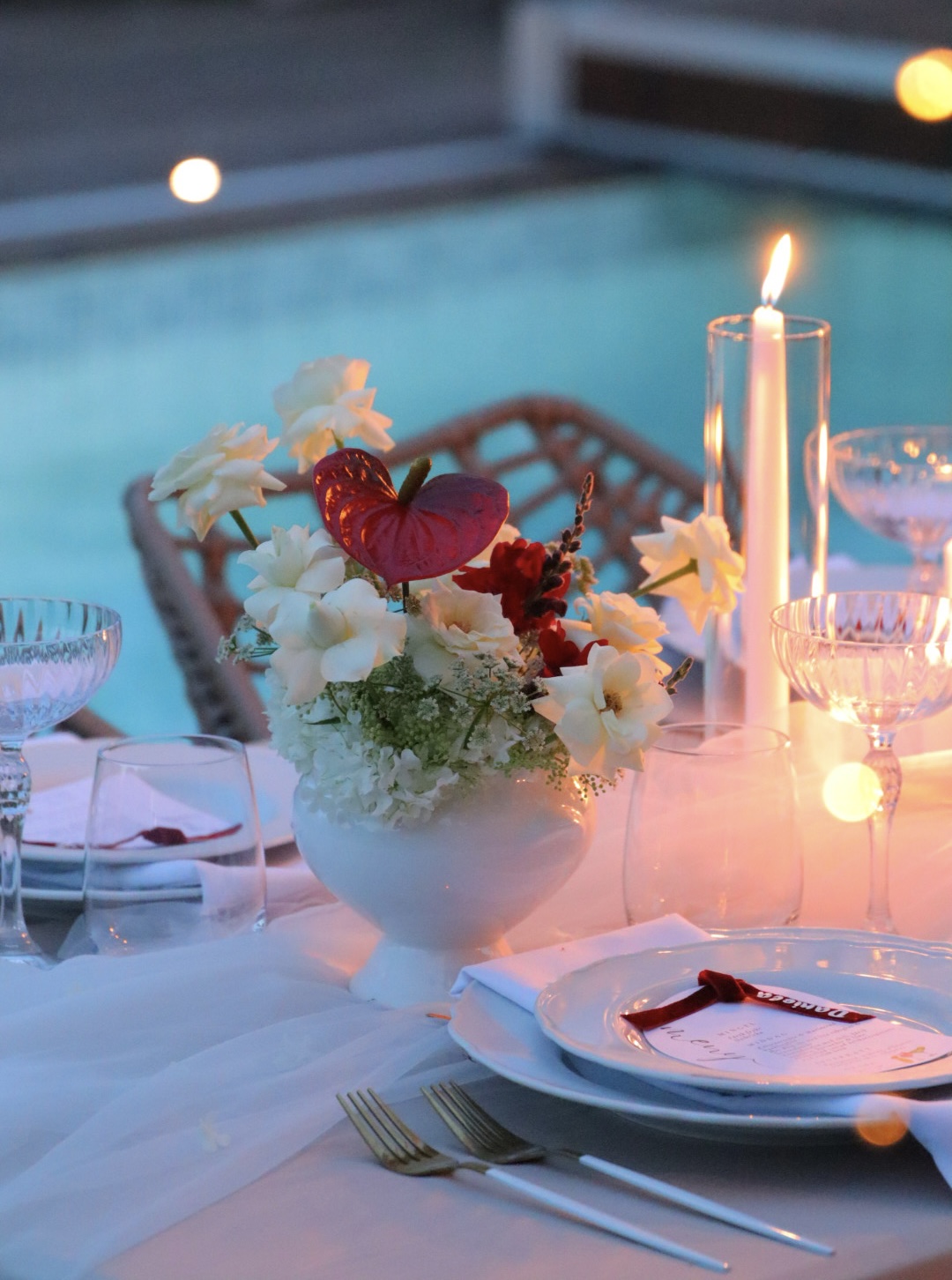 Table setting with a floral centerpiece, lit candle, and glassware near a swimming pool at night.