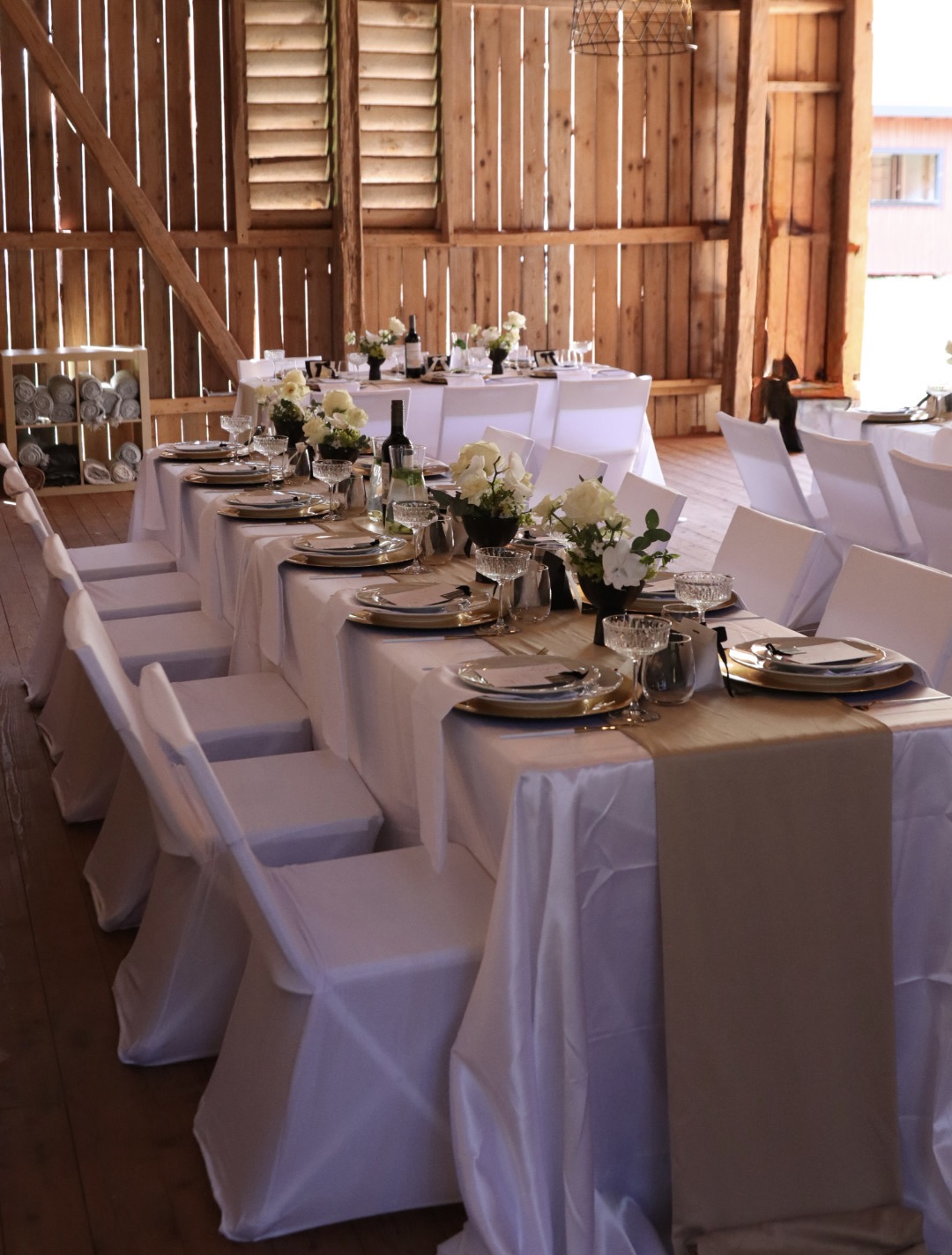Long dining table with white tablecloth, floral centerpieces, and place settings in a rustic wooden barn interior.
