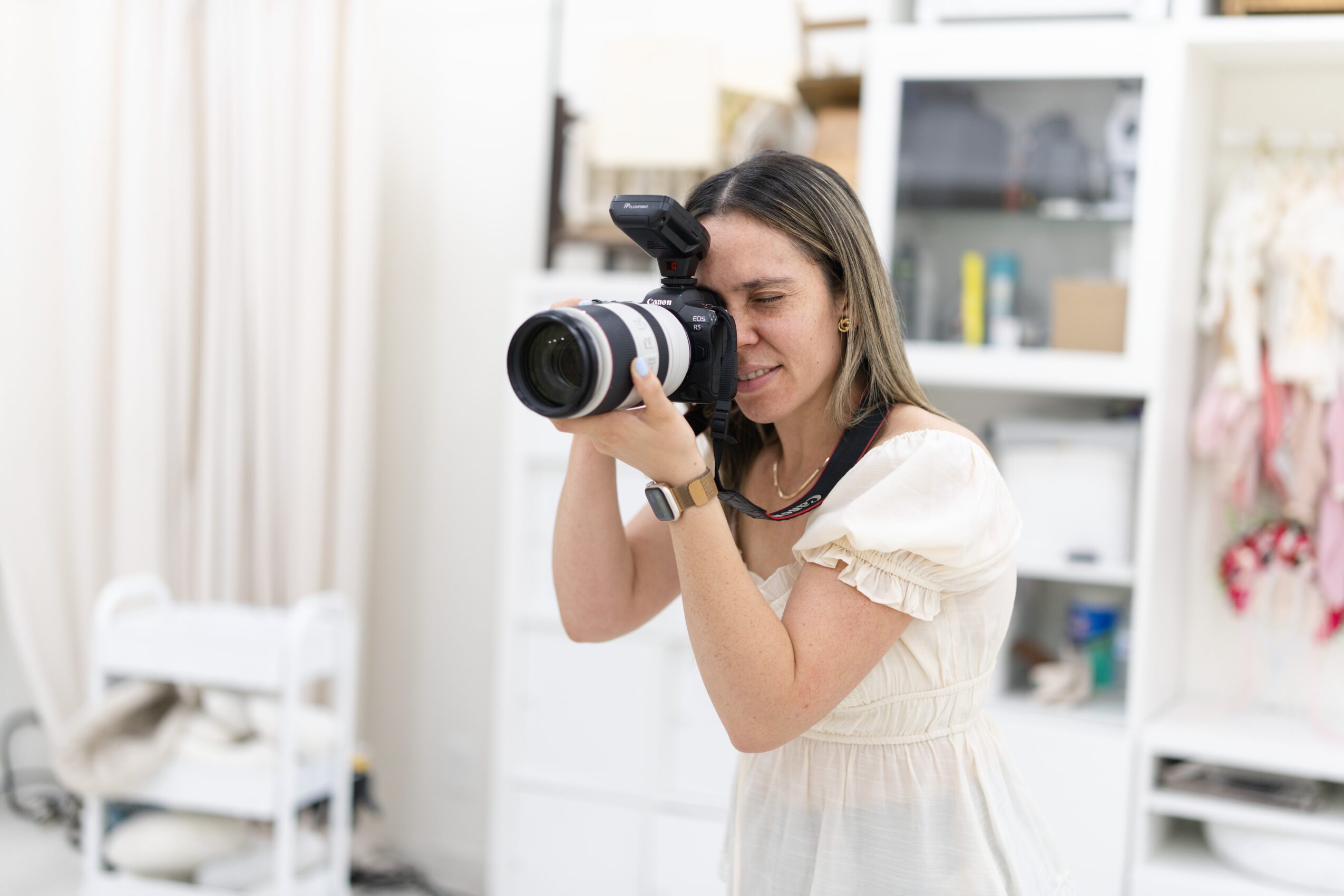 Woman taking a photo with a camera indoors, smiling, in a room with shelves and curtains