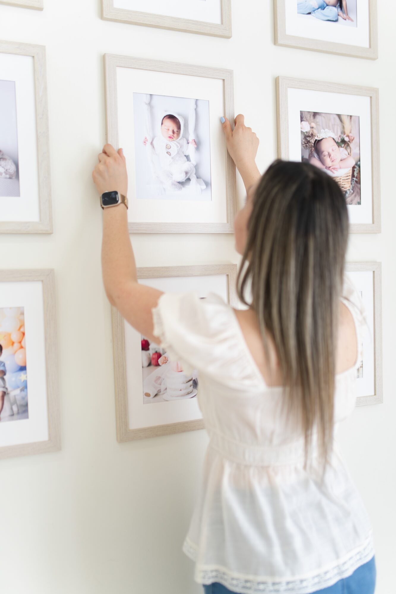 Person with long hair and white top adjusting a framed photo on a wall with multiple picture frames, facing away from the camera.