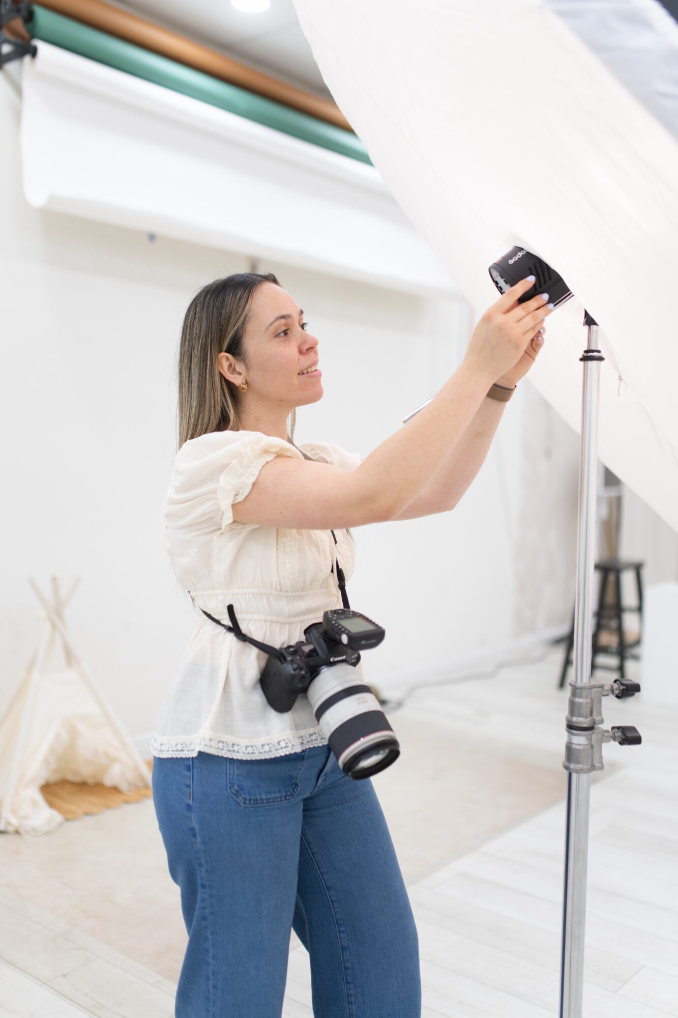 Woman holding a smartphone and camera in a photography studio with a white backdrop, adjusting lighting equipment.