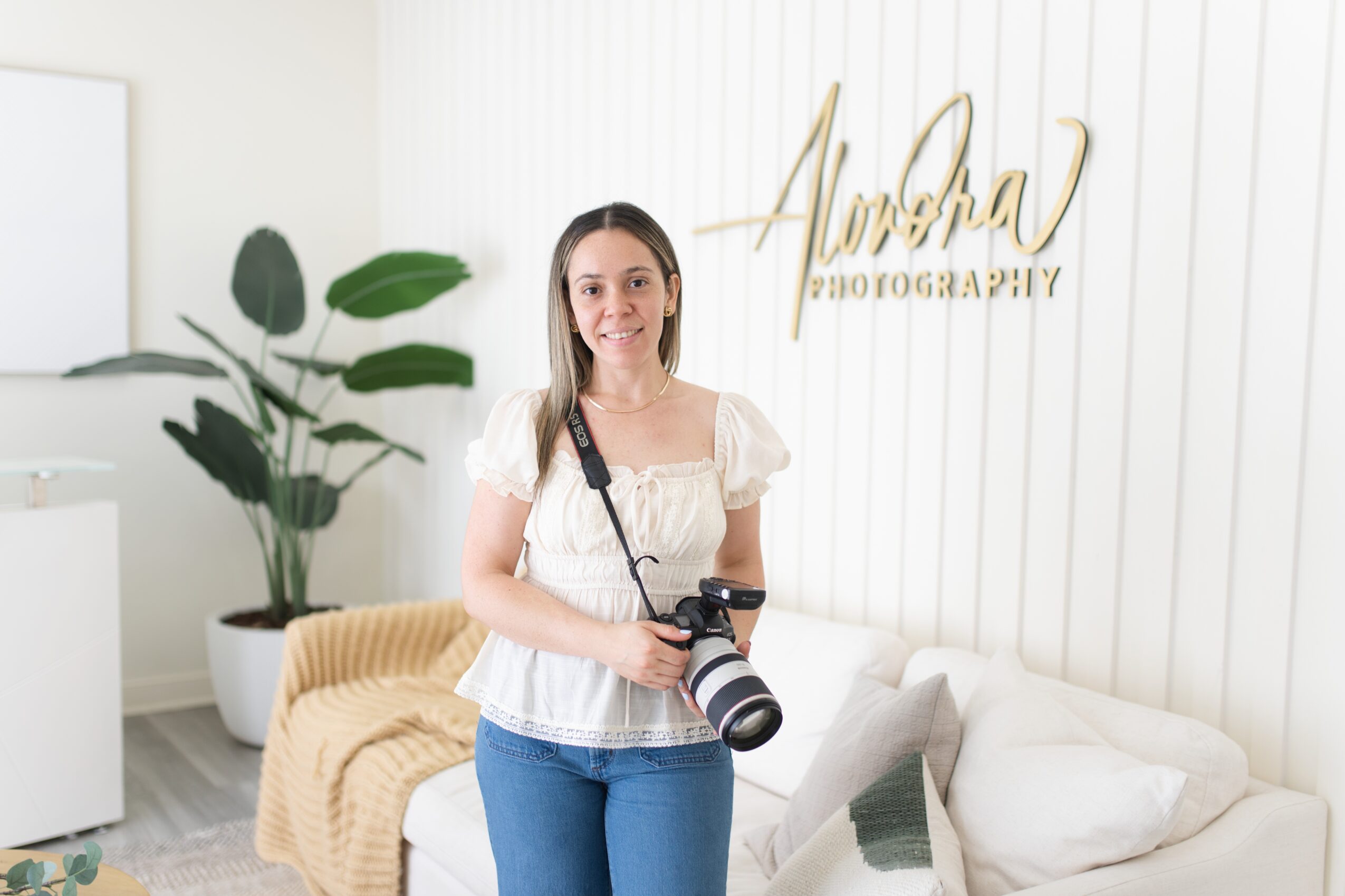 Young woman holding a camera with a strap, standing in a photography studio with a white wall and a plant.