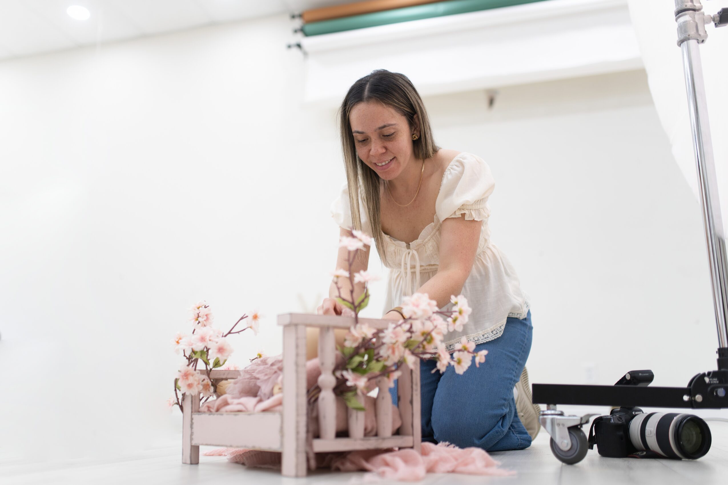 Woman kneeling next to a small bed with flowers, smiling, in a bright room with a camera on a tripod nearby.