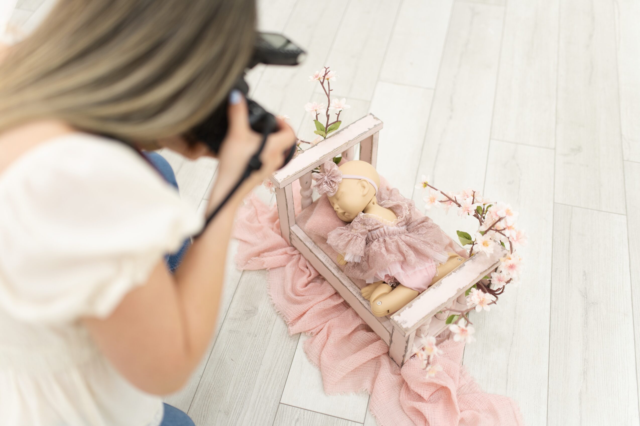 Person taking a photo of a doll in a small bed with pink fabric and flowers, on a light-colored floor.