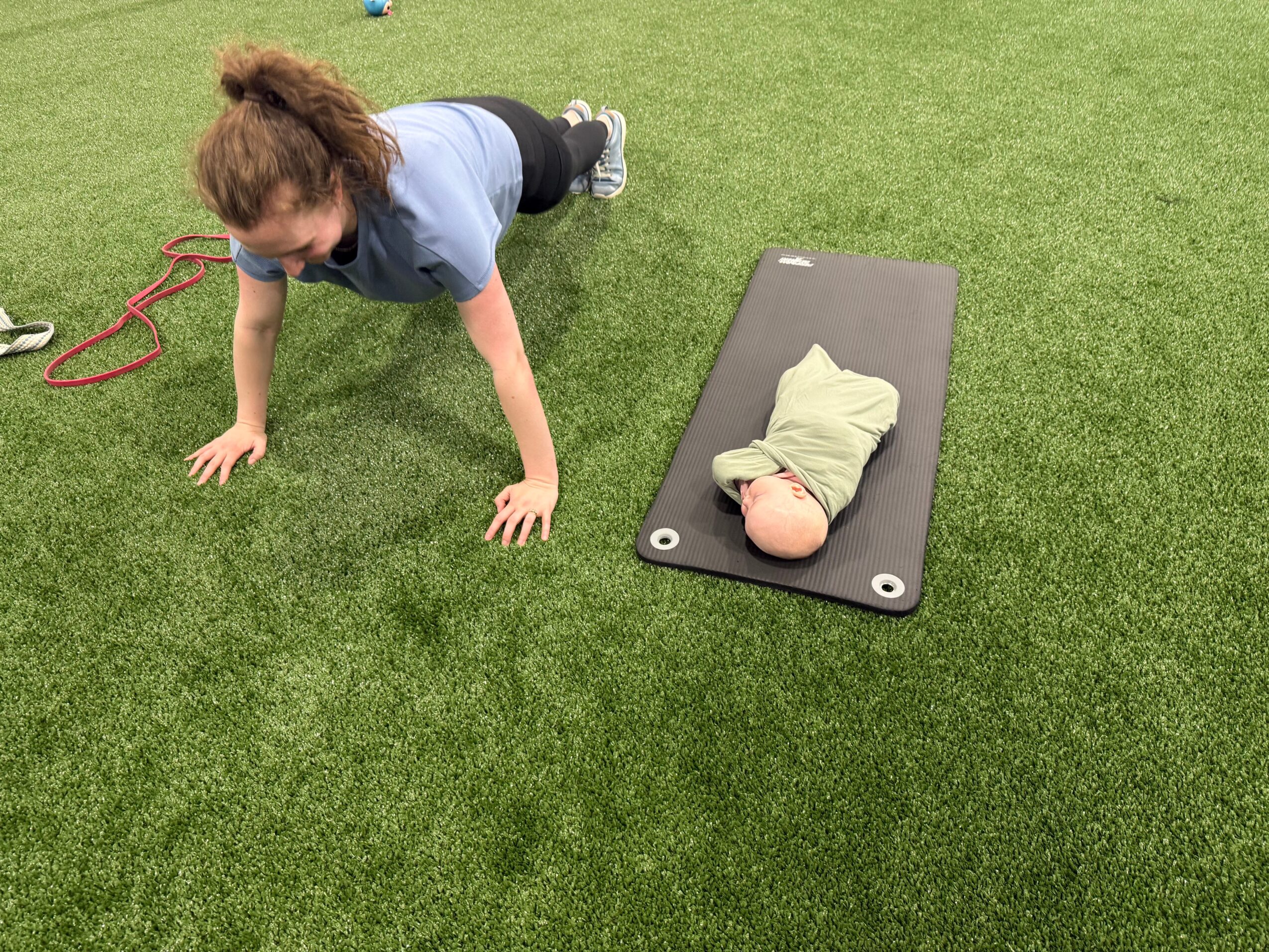 Child doing push-up on grass next to a baby wrapped in a blanket on a mat.