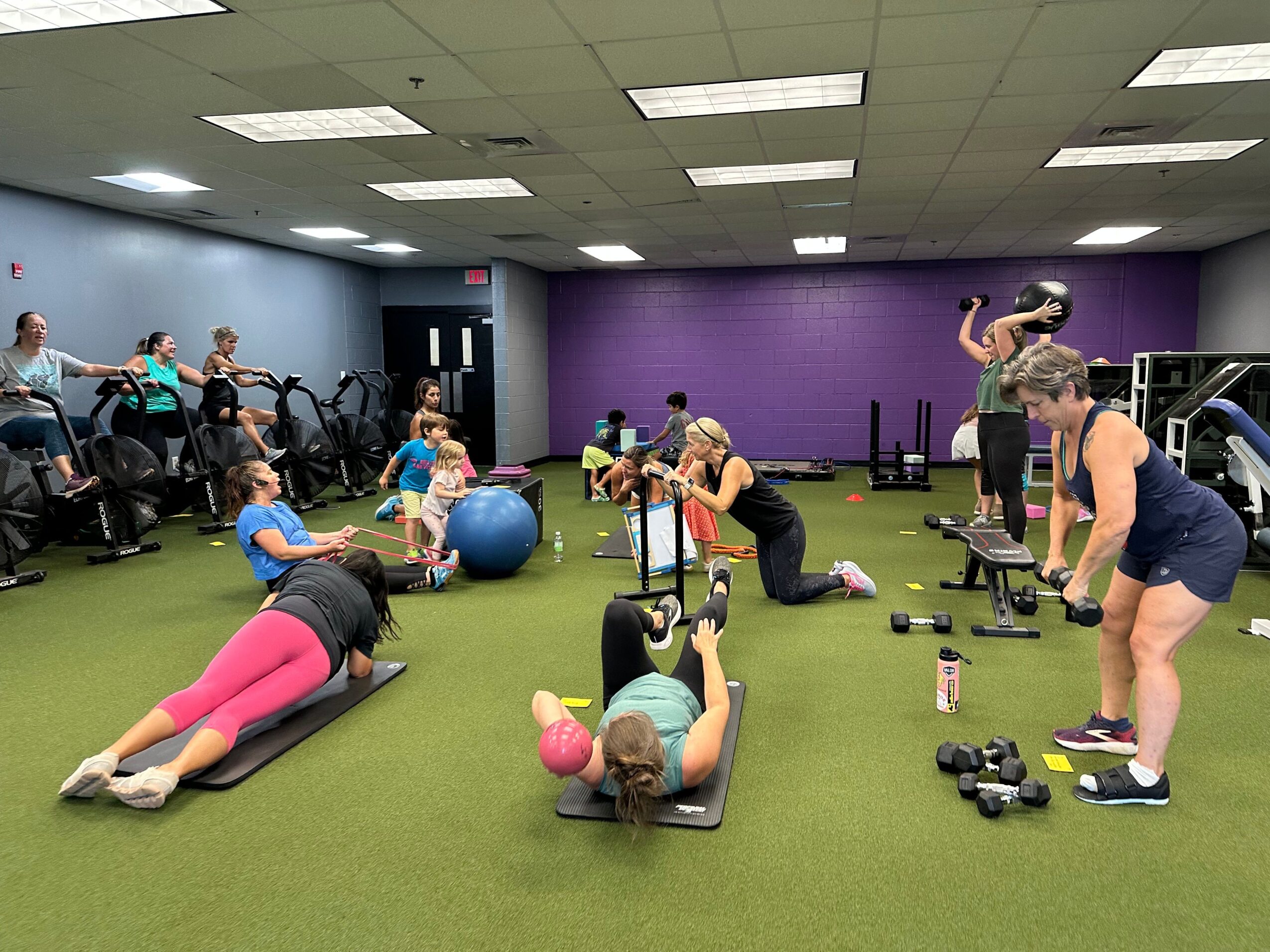 People exercising on mats and equipment in a gym with purple and gray walls, green floor, and bright lighting.