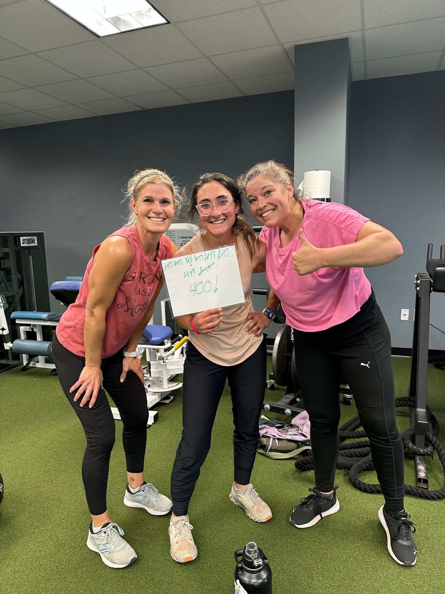 Three women in athletic clothing holding a sign, standing on green artificial turf in a gym or fitness center.