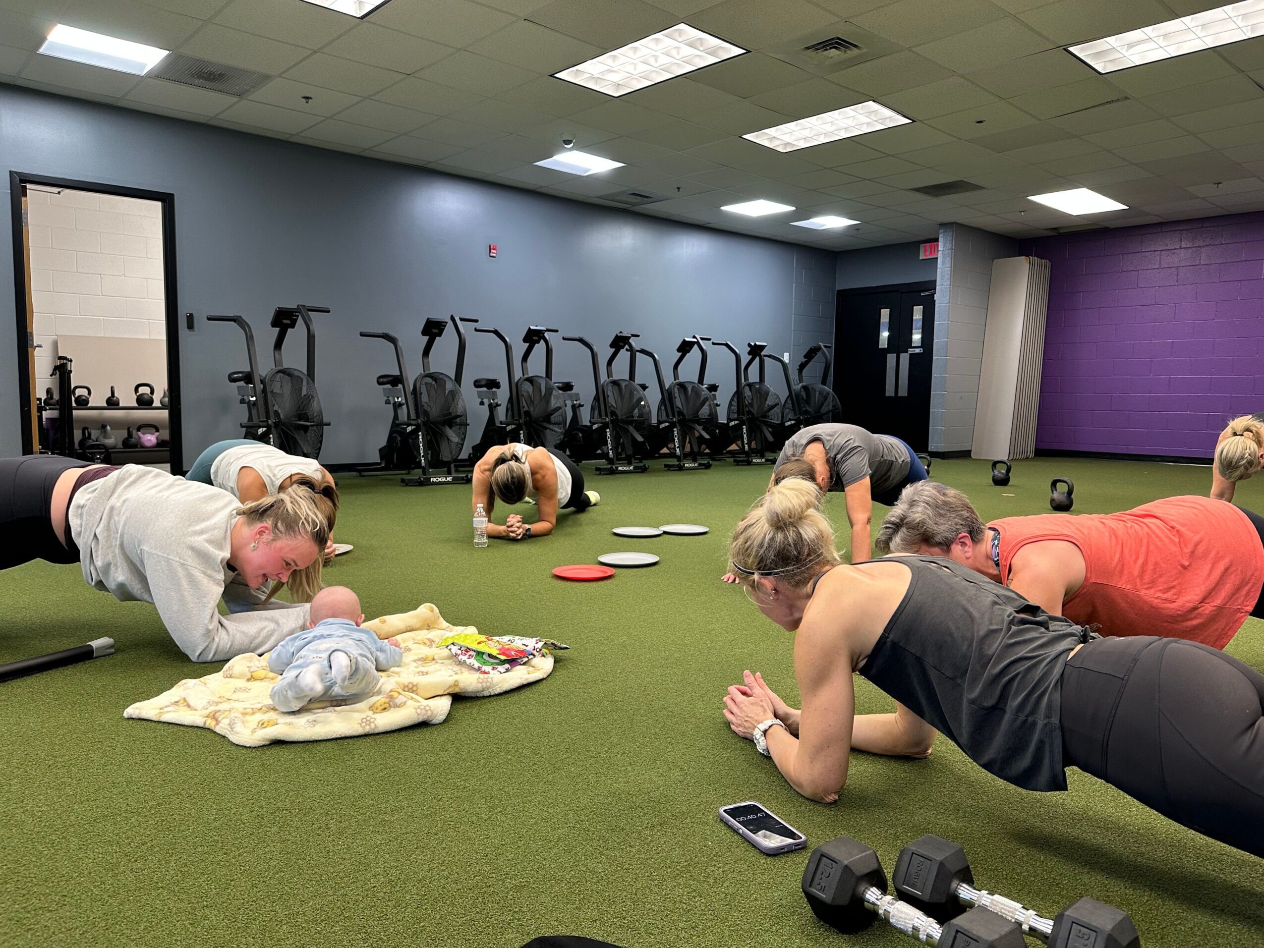 Group of children and an adult sitting on green carpeted floor in a room with exercise equipment, engaging in a discussion.