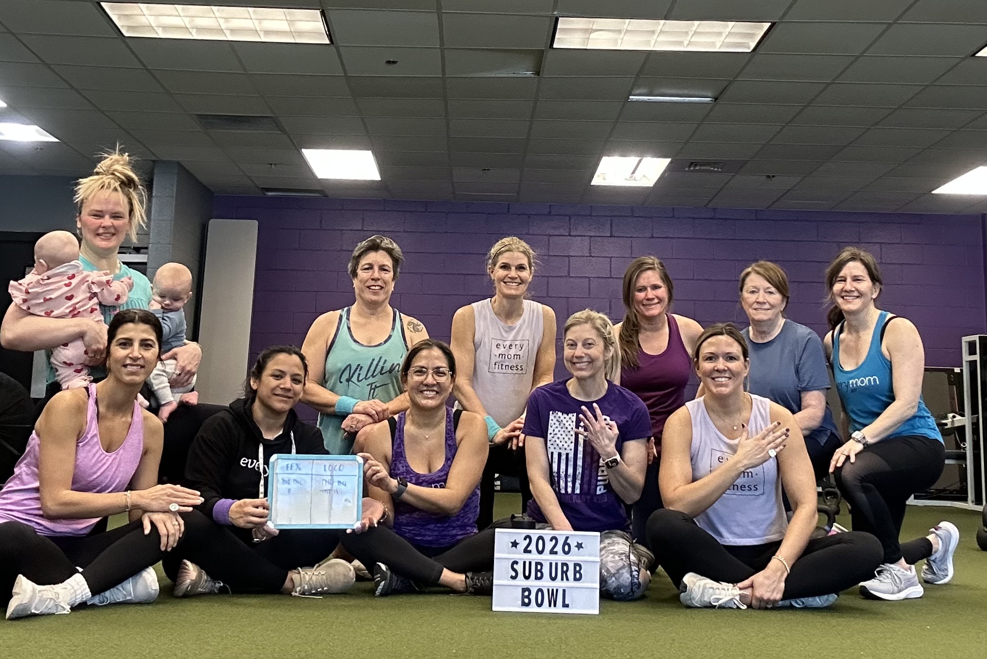 Group of women and children in a gym, some sitting and some standing, smiling, with a purple wall in the background.
