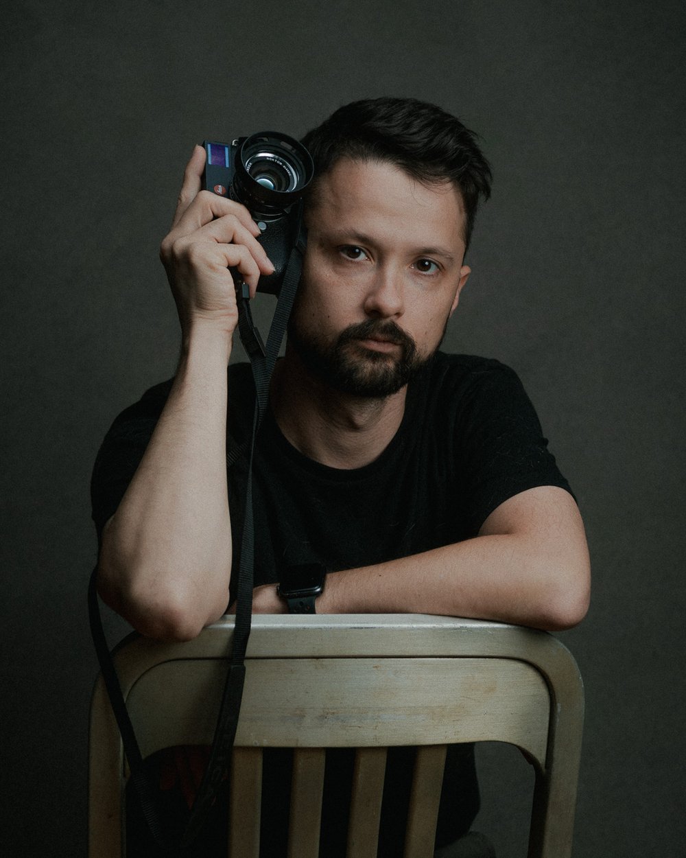 Man with dark hair and beard holding a camera, resting his arm on a chair, against a dark background.