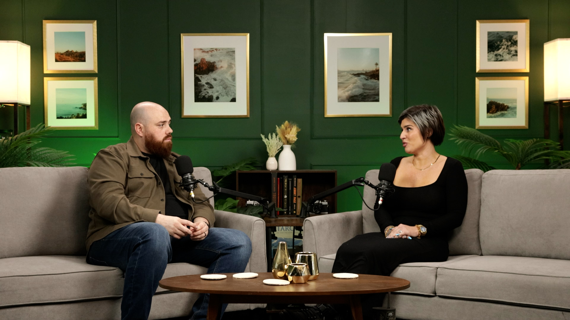 Two people sit on sofas in a room with green walls and framed pictures, having a conversation with microphones.