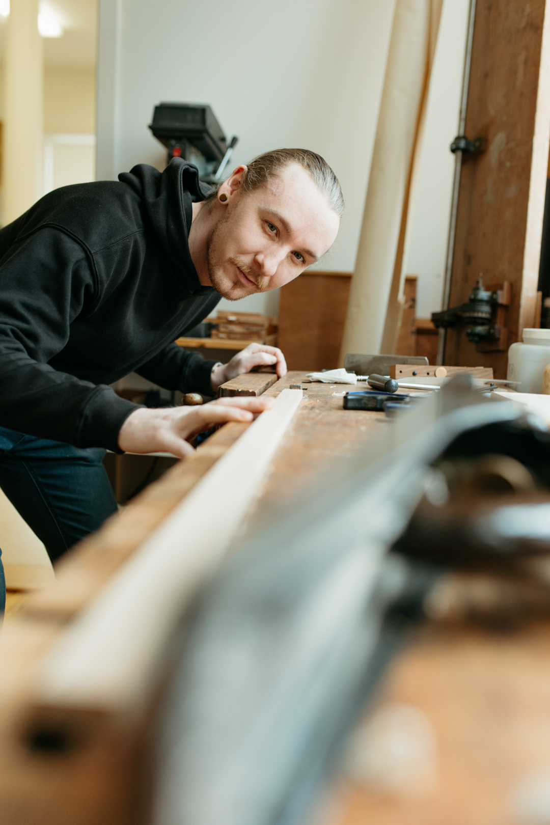 Person with long hair tied back working at a wooden table in a workshop, surrounded by tools and equipment.