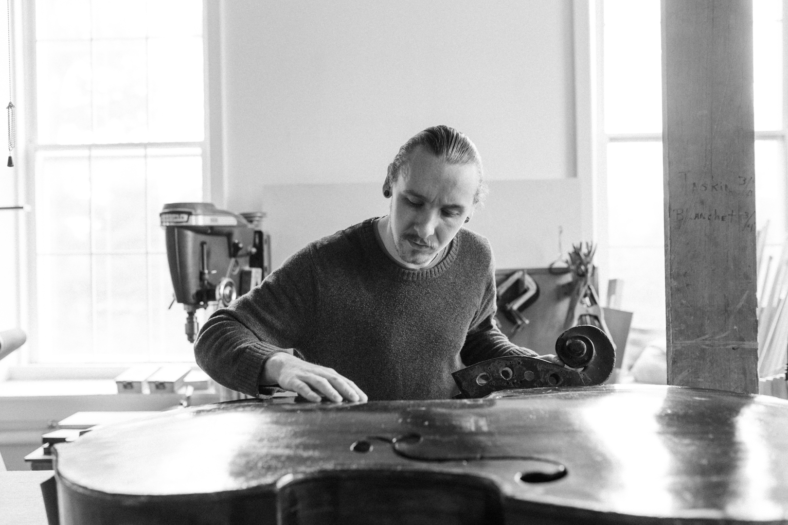 Person working on a large, curved wooden object in a bright workshop with windows and tools nearby.