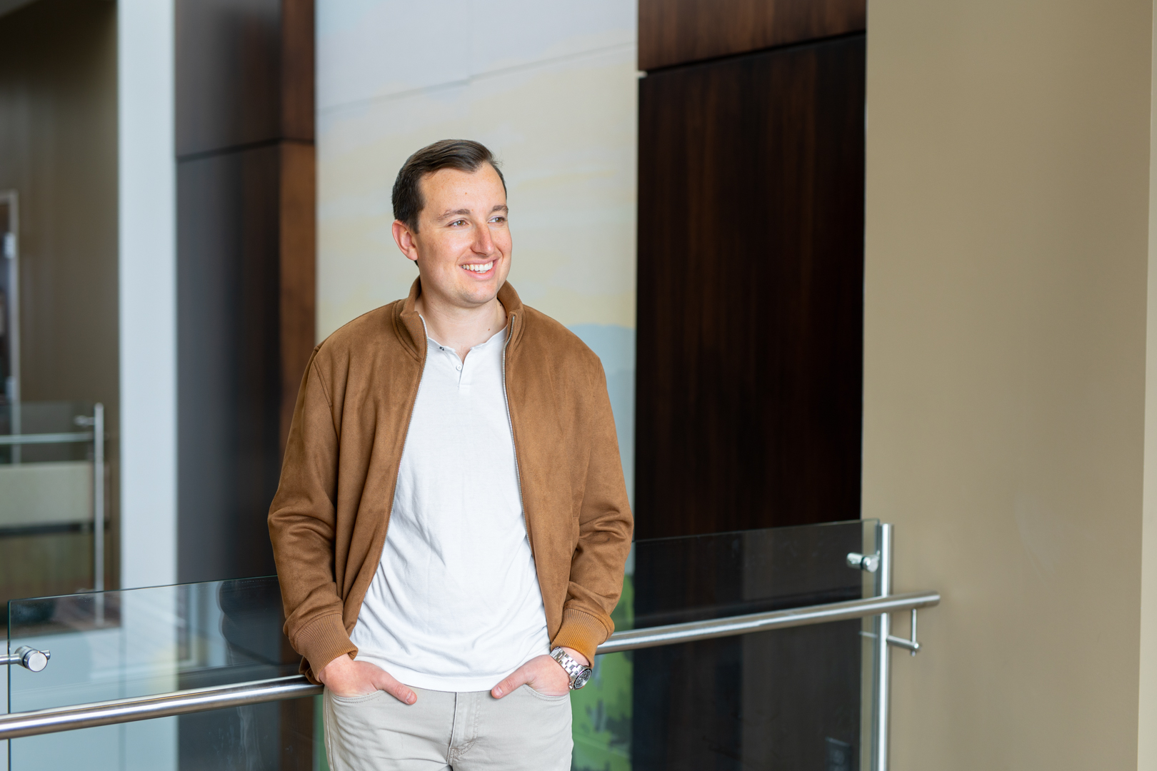 Man smiling, standing indoors near a glass railing, wearing a brown jacket and white shirt.