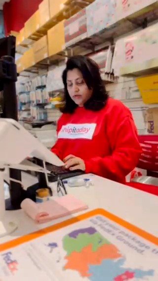 Woman with dark hair wearing a red sweatshirt working at a checkout counter in a store, shelves with boxes behind her.