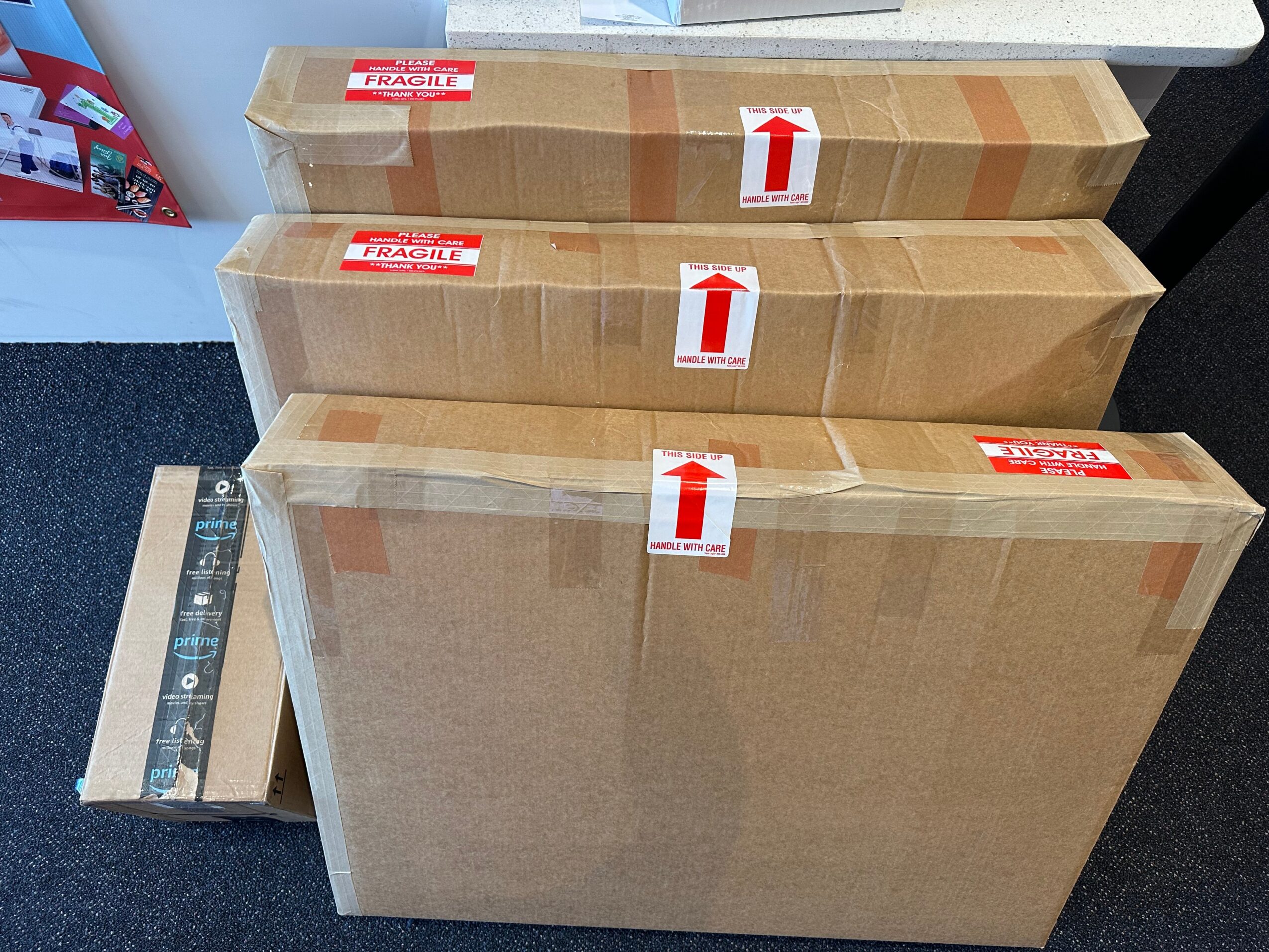 Three large cardboard boxes stacked on a table, with red and white labels and arrows, in a room with a ruler nearby.