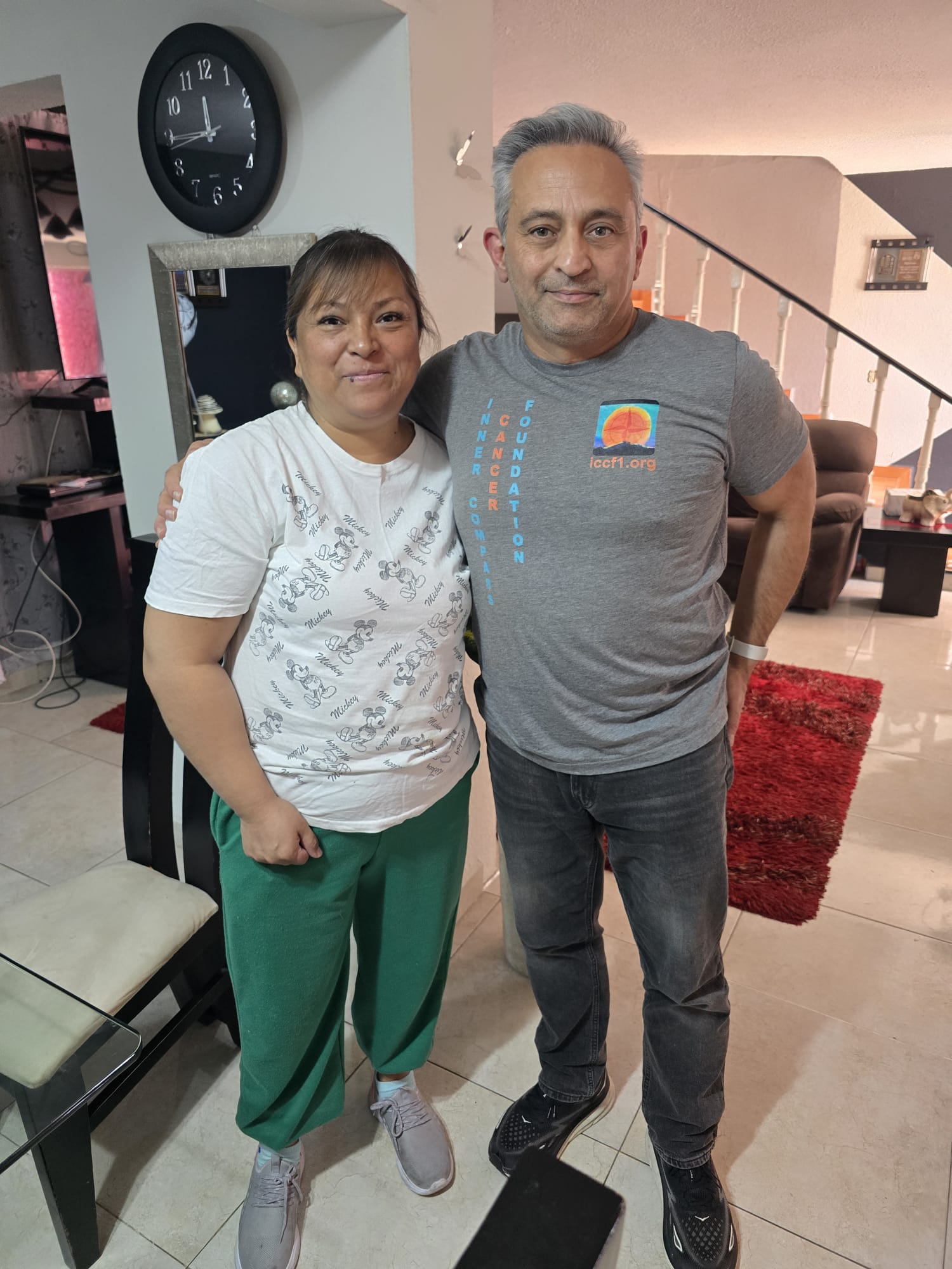 Two people standing indoors, a woman in white and green clothing and a man in a gray t-shirt and jeans, smiling at the camera.