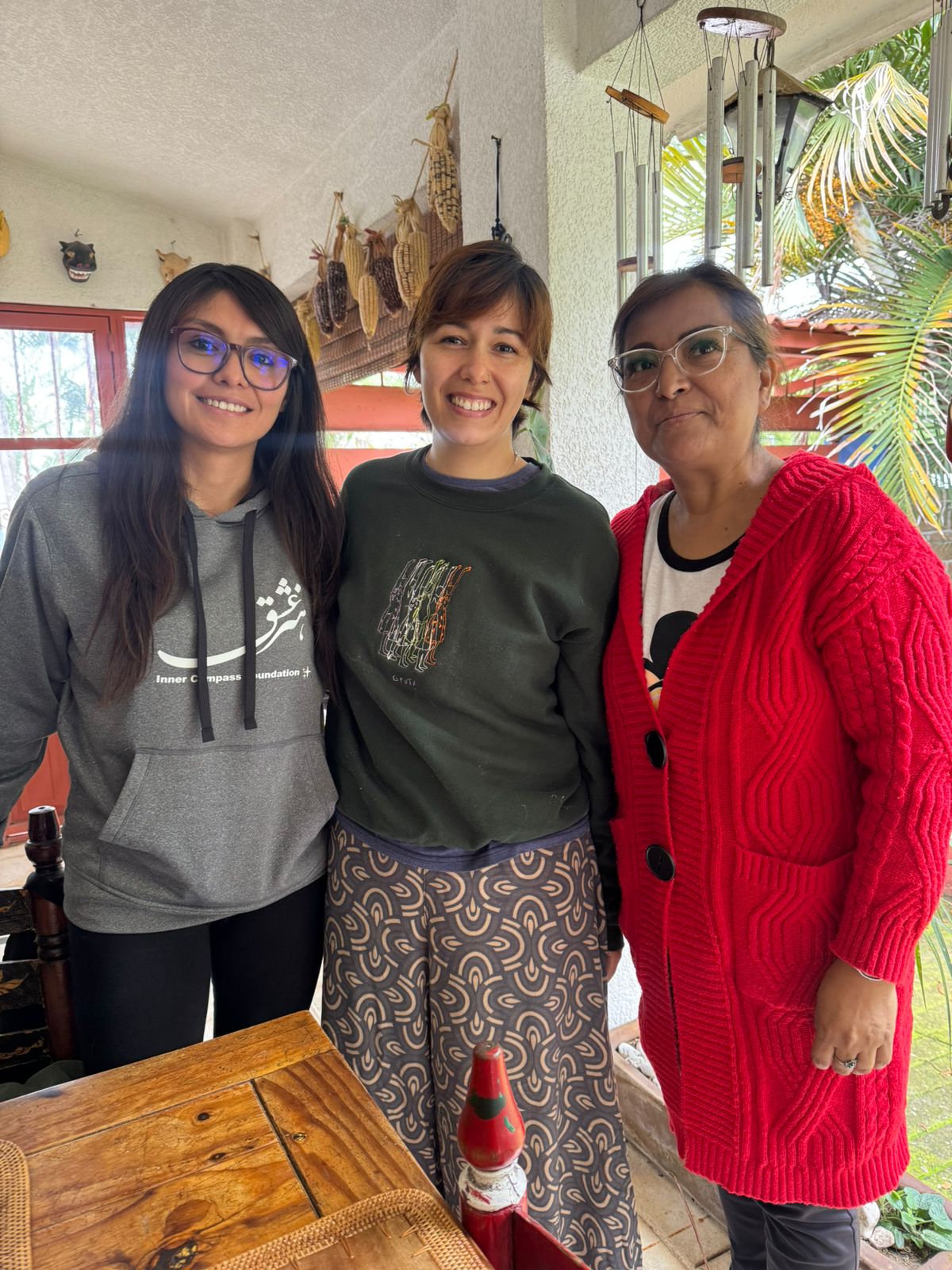 Three women standing indoors, smiling, with plants and a window in the background.