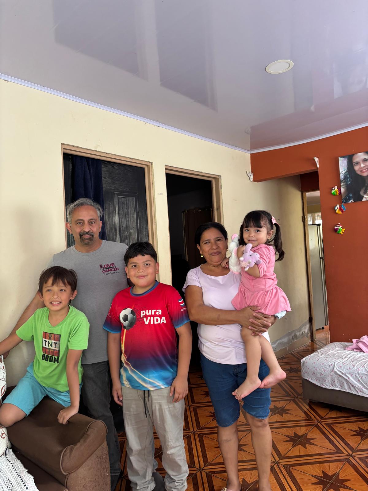 Group of five people, including two children, standing and sitting indoors, smiling, with a bed and colorful wall in background.