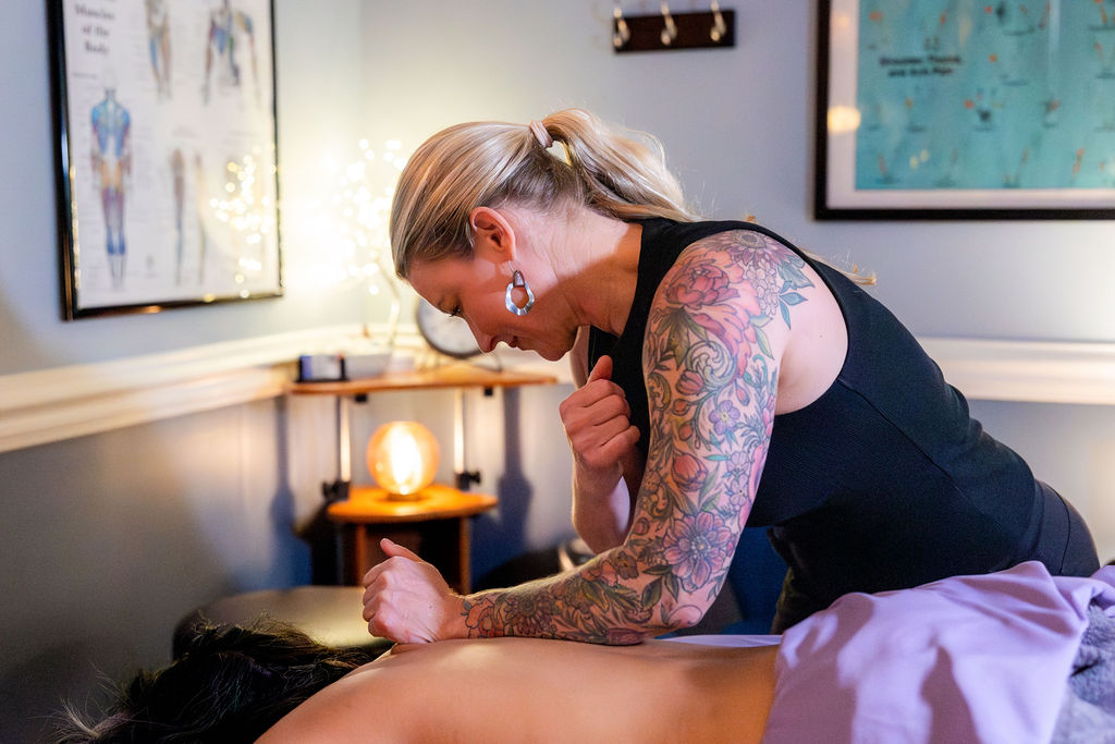 Woman with tattoos on her arm leaning over a person lying down in a room with framed artwork and a lamp, touching her shoulder.