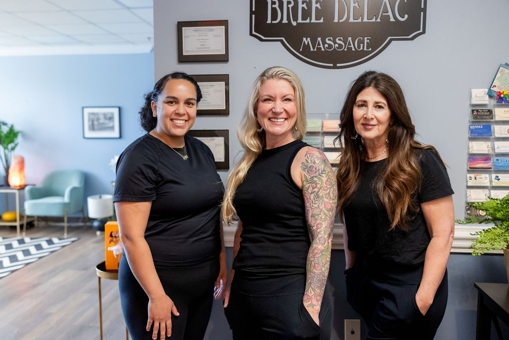 Three women standing inside a massage therapy clinic, smiling, with a sign reading 'Bree Bellac Massage' behind them.