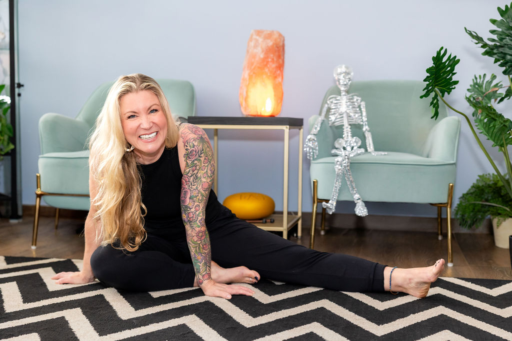 Woman with long blonde hair and tattoos smiling on the floor in a modern living room, with a salt lamp and skeleton model behind her.