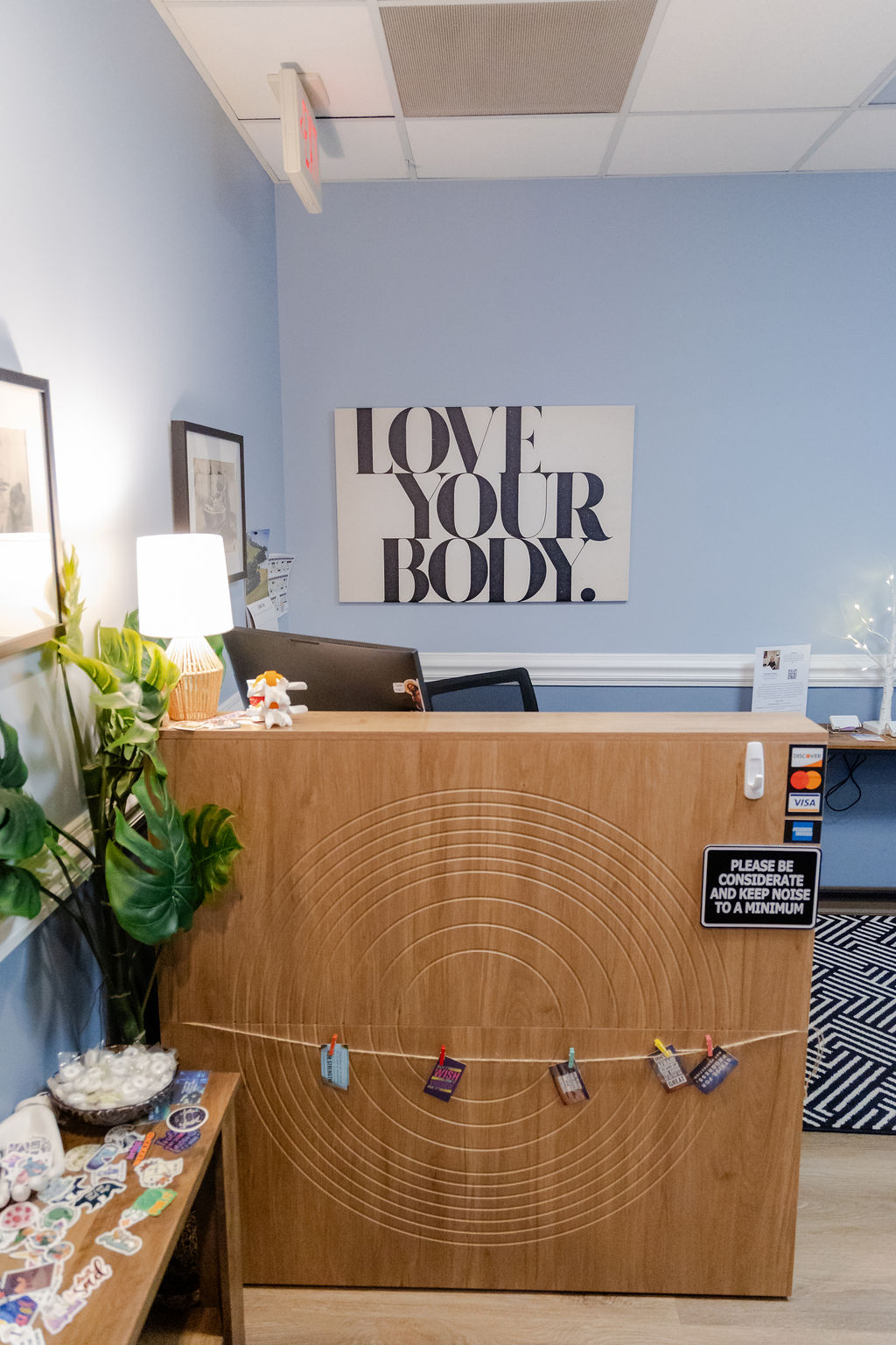 Reception desk with a sign reading 'LOVE YOUR BODY' on a blue wall, plants, and office supplies visible.