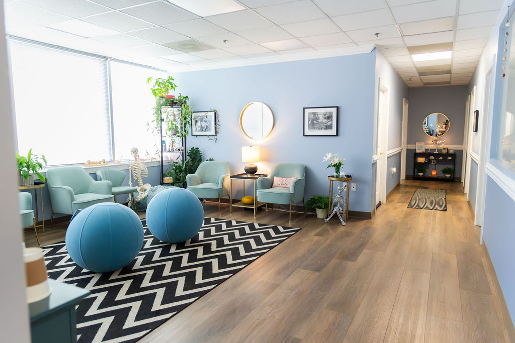 Bright waiting area with blue walls, seating, plants, and a black-and-white zigzag rug, with a hallway on the right.