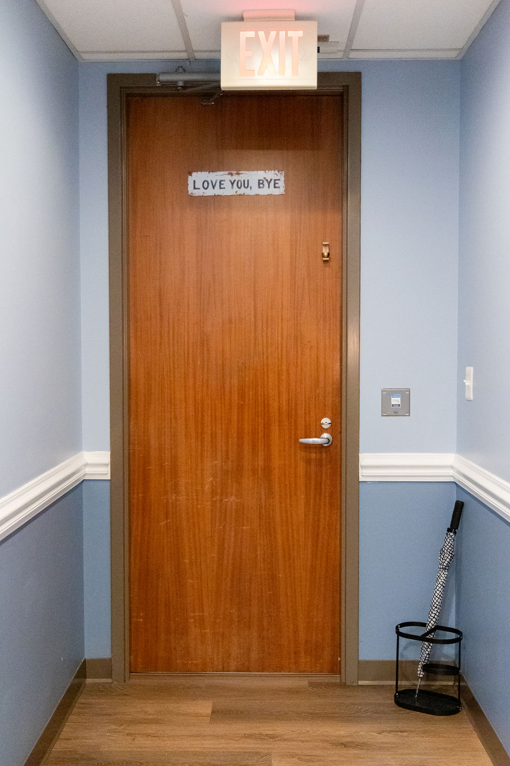 Wooden door with a sign that says 'LOVE YOU, BYE' in a hallway with blue walls and a chair on the right.