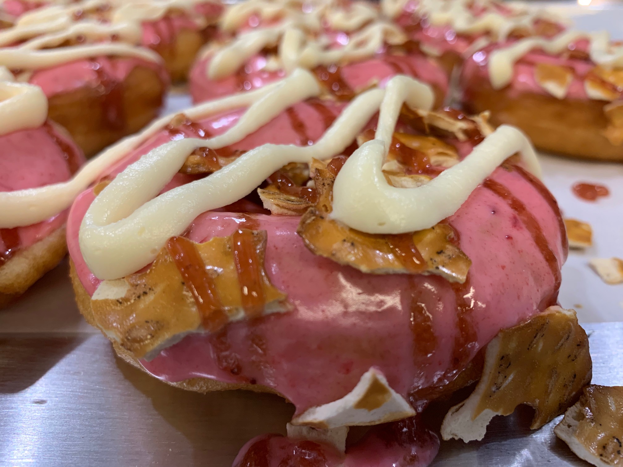Close-up of a decorated donut with pink glaze, white icing, bacon pieces, and caramel drizzle, on a tray.
