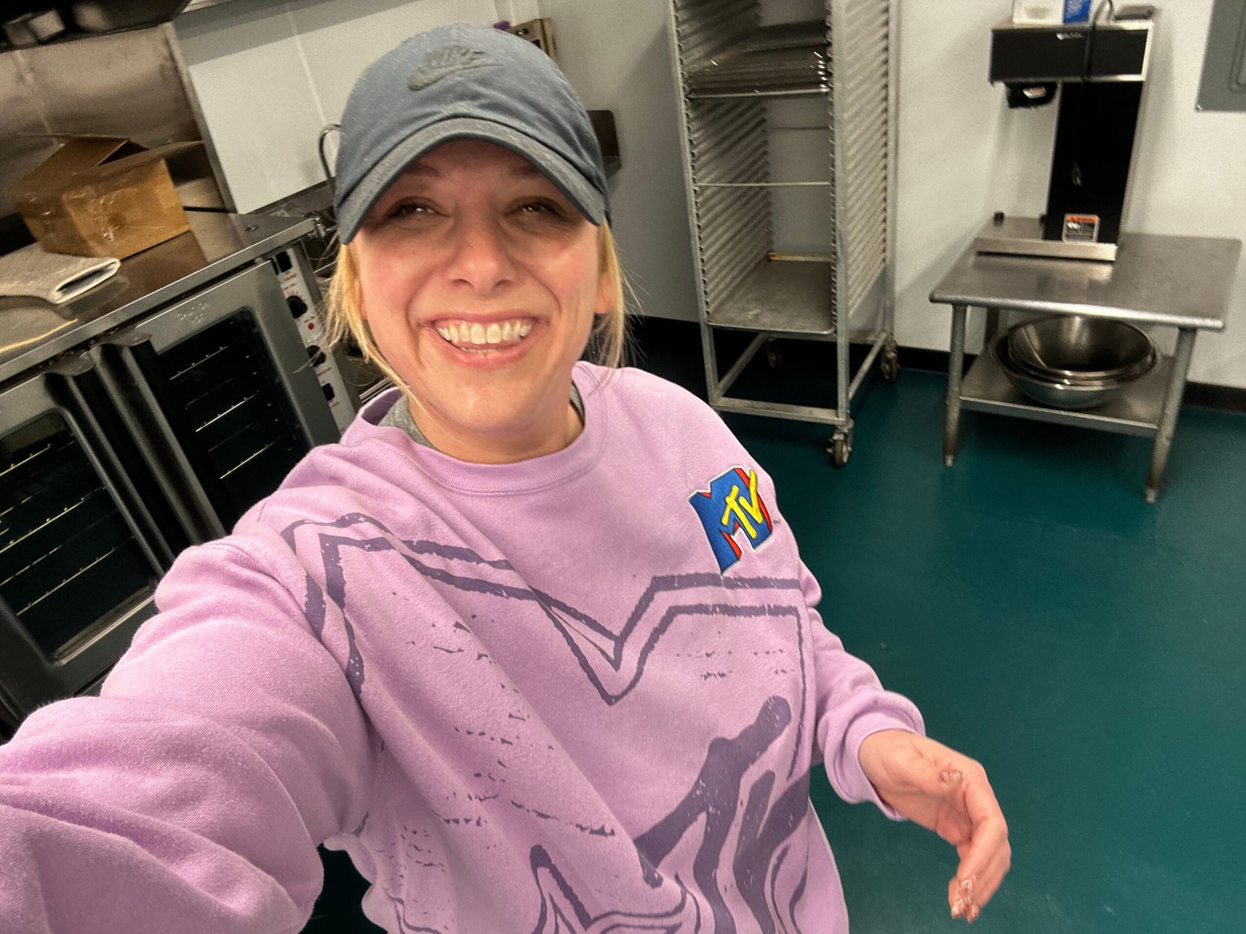 Person smiling, wearing a gray head covering and purple hoodie, in a kitchen with teal wall and metal shelves.