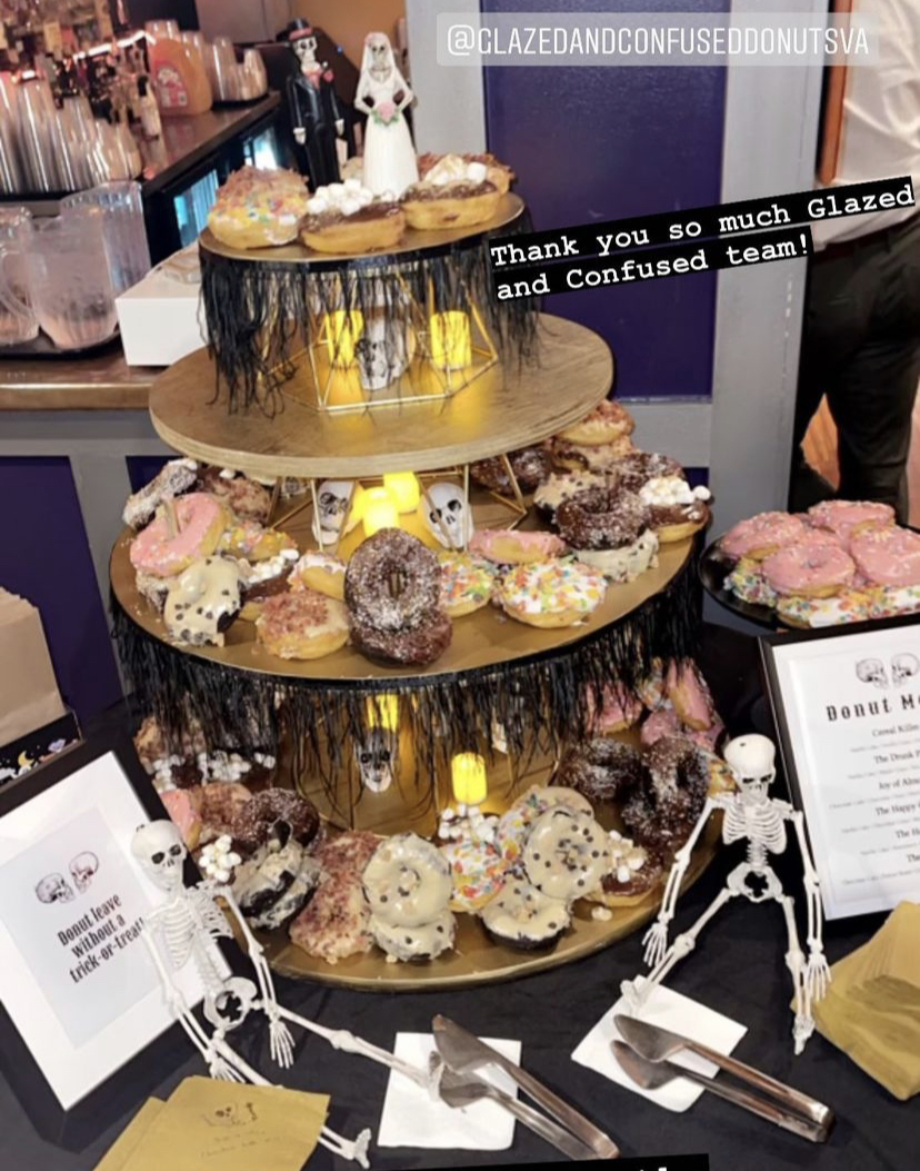 Three-tiered dessert stand with assorted donuts and pastries, decorated with small skeleton figures, in a cafe setting. Text overlay thanks Glazed and Confused team.