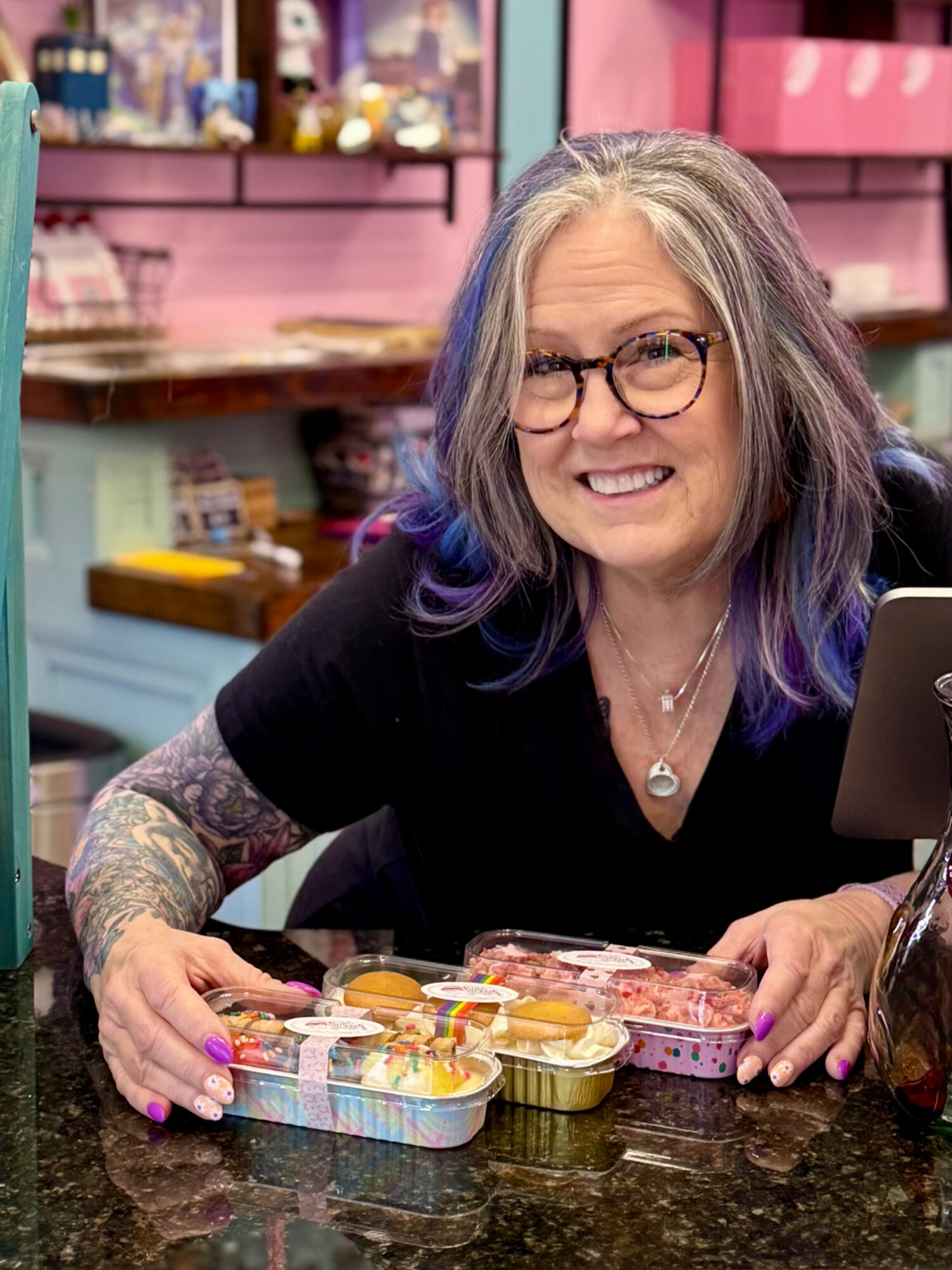 Woman with glasses and gray hair with purple streaks smiling at a counter with colorful containers.