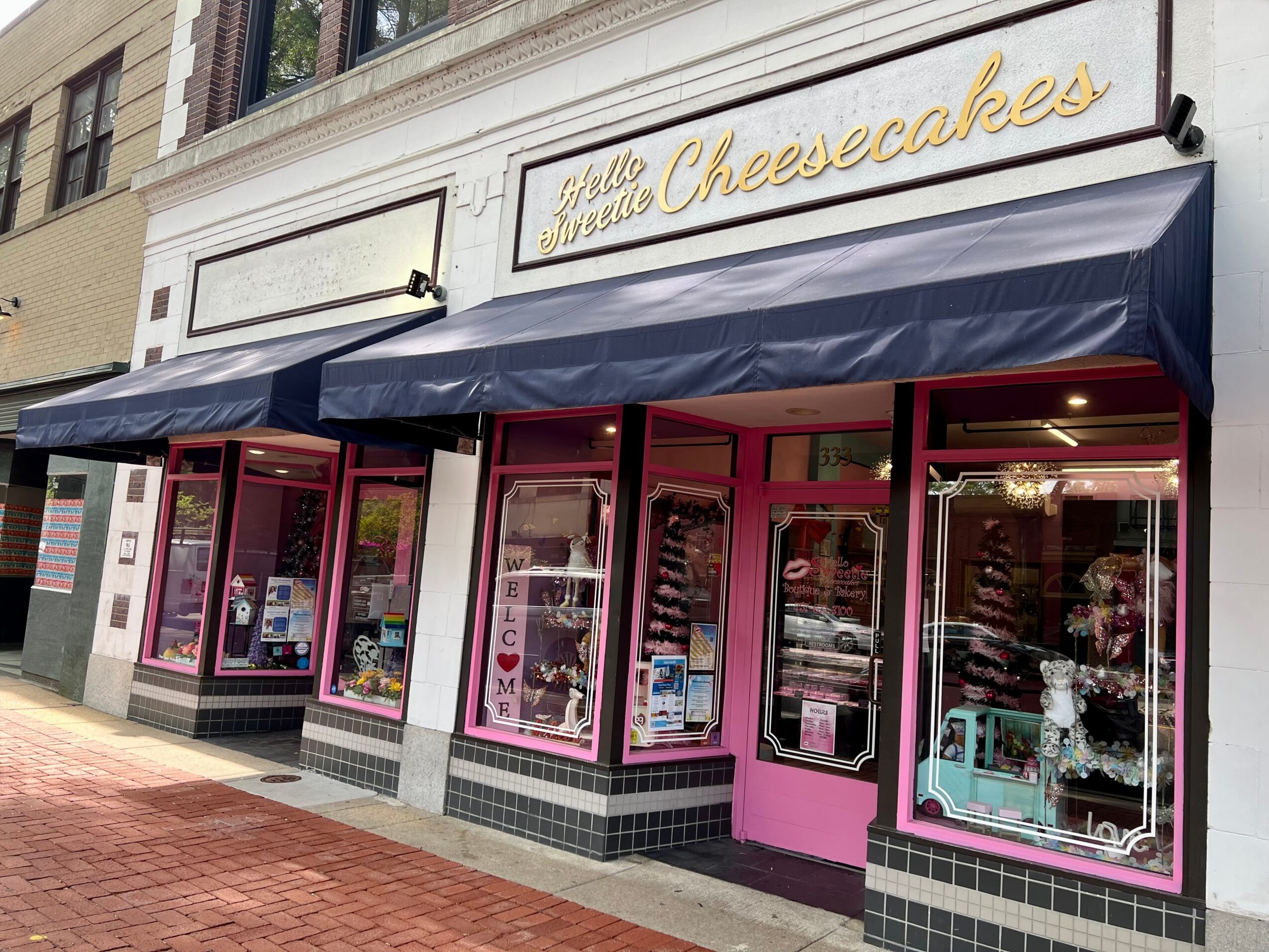 Storefront with pink window frames and a blue awning, signage reads 'Hello Miss Cheesecakes'.