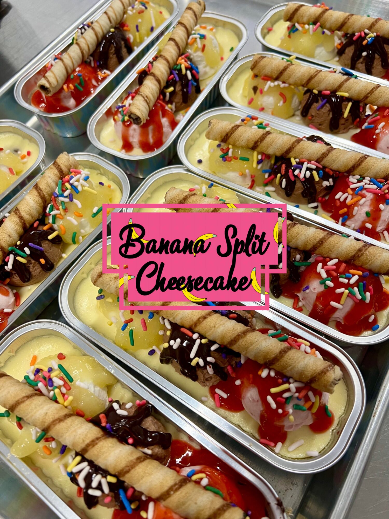 Multiple banana split cheesecakes in metal trays topped with whipped cream, chocolate, and colorful sprinkles, with wafer rolls on top.