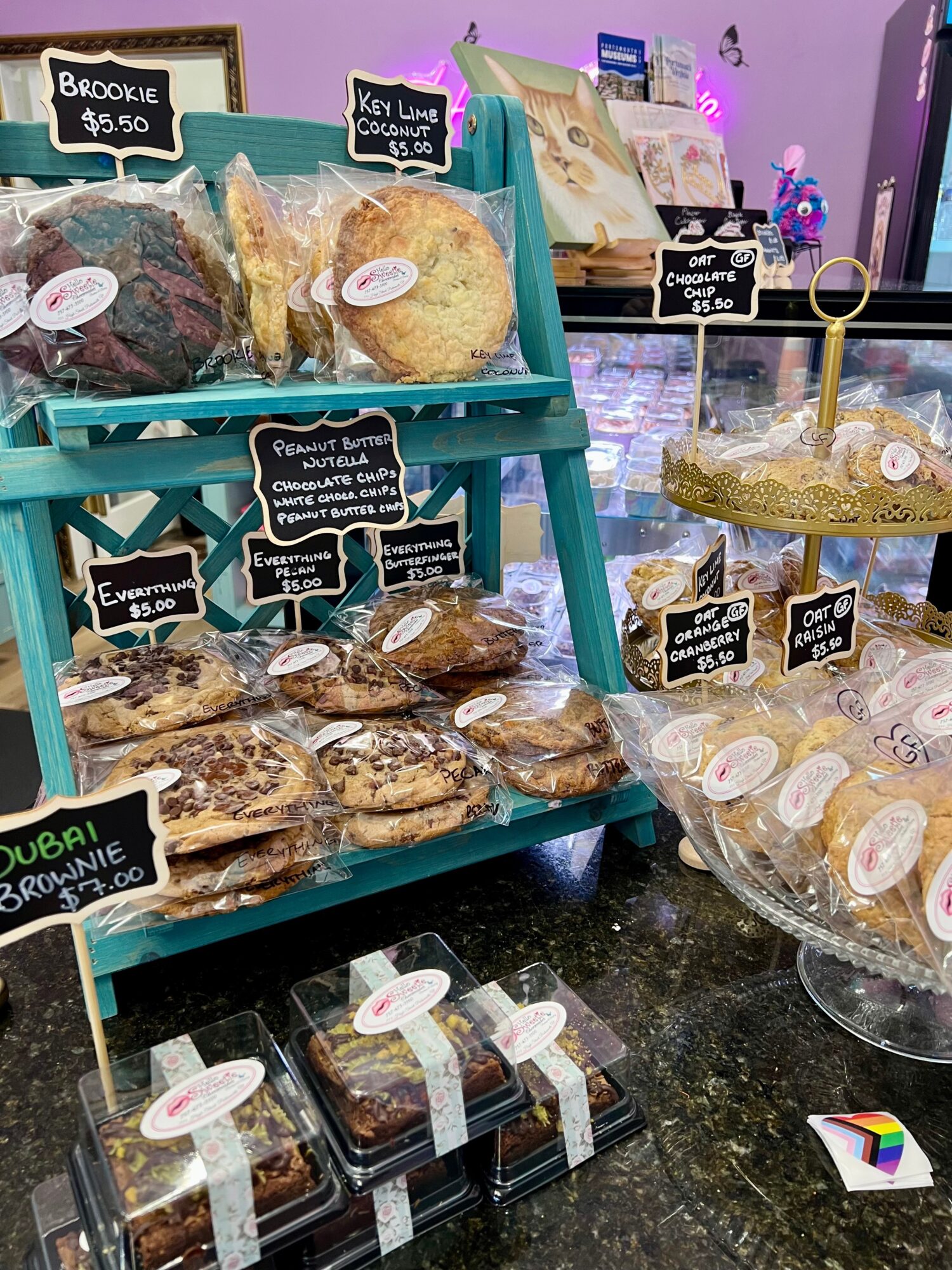 Display of baked goods with signs, including cookies, brownies, and muffins, on a table with purple background.
