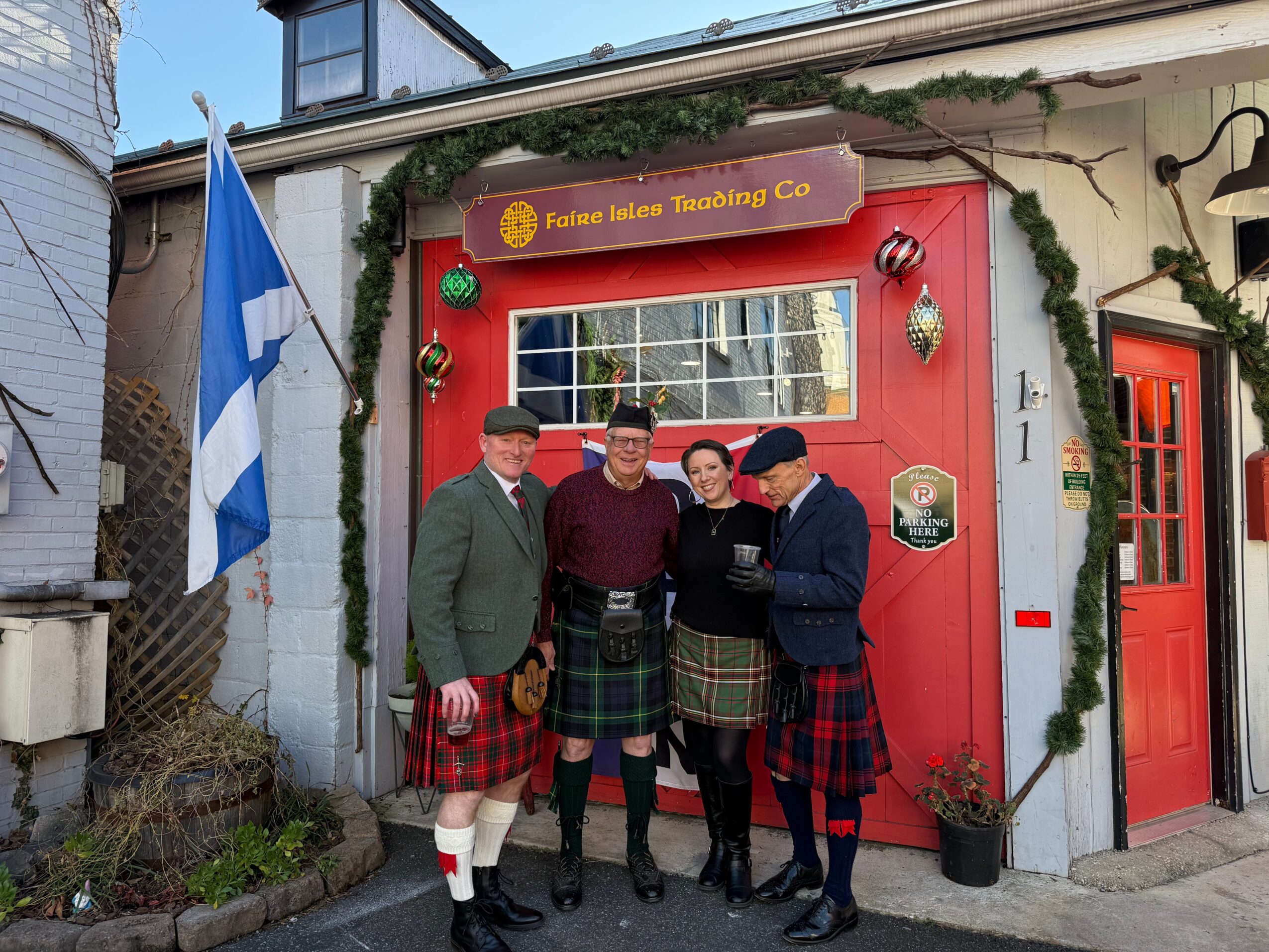 Four people in traditional Scottish attire stand outside a building decorated for St. Patrick's Day, with a flag and greenery.