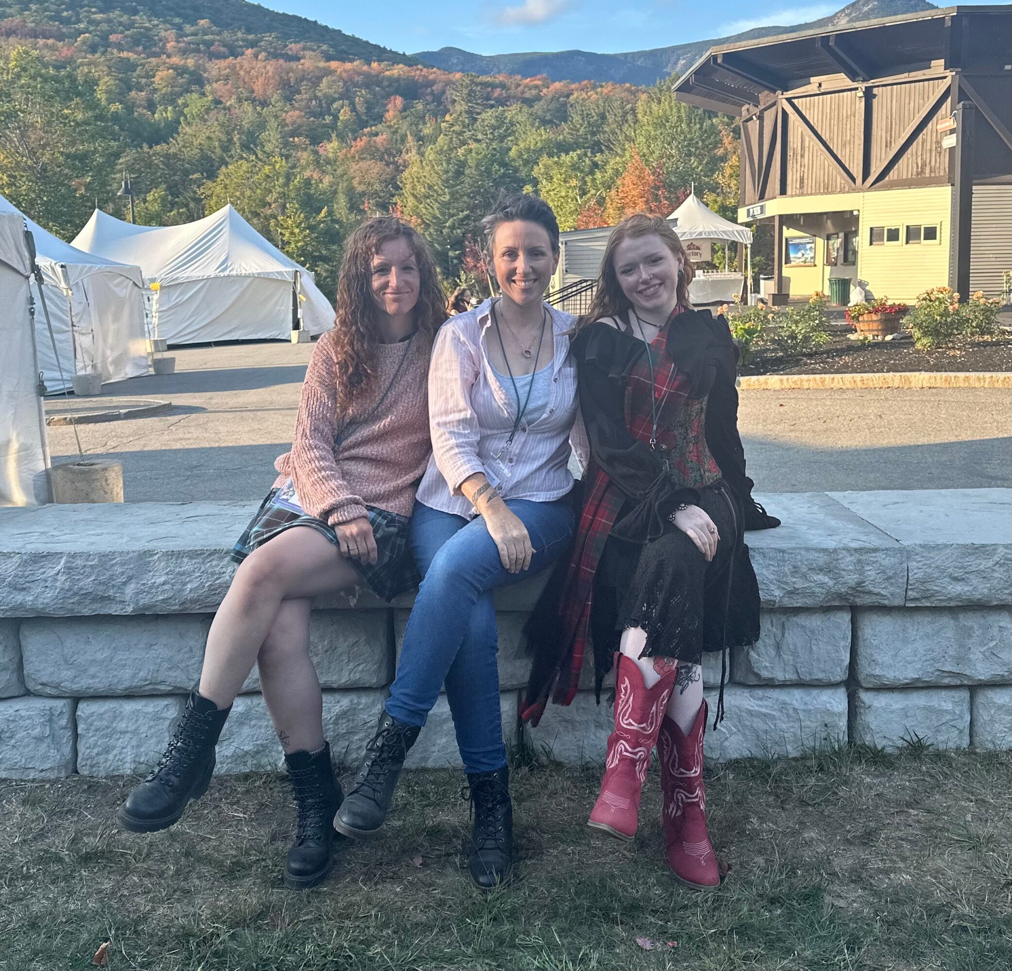 Three women sitting on a stone wall outdoors with tents and a building in the background, trees, and mountains.