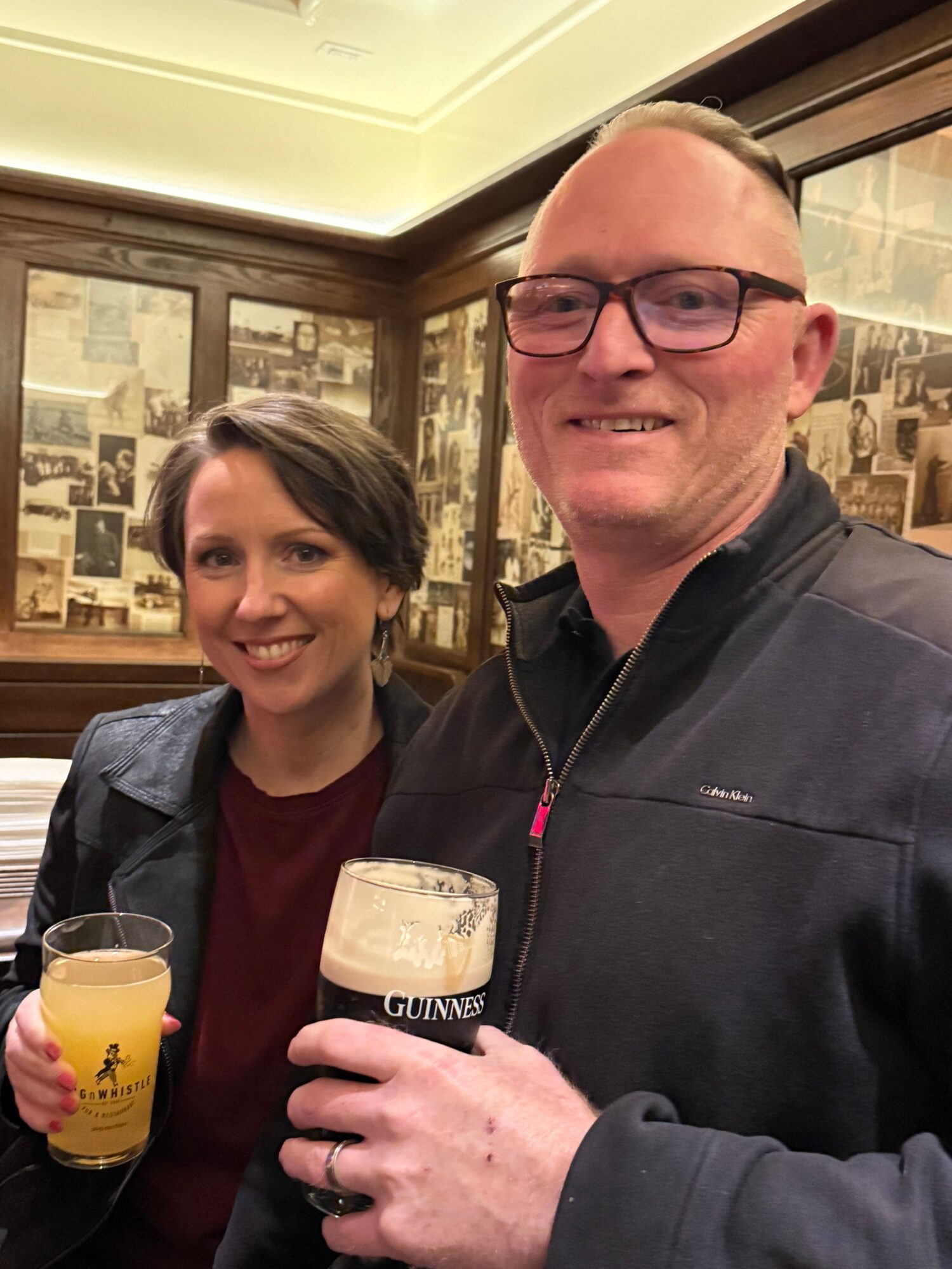 Two people smiling at a bar, one holding a Guinness beer and the other holding a yellow drink, with a background of framed pictures.