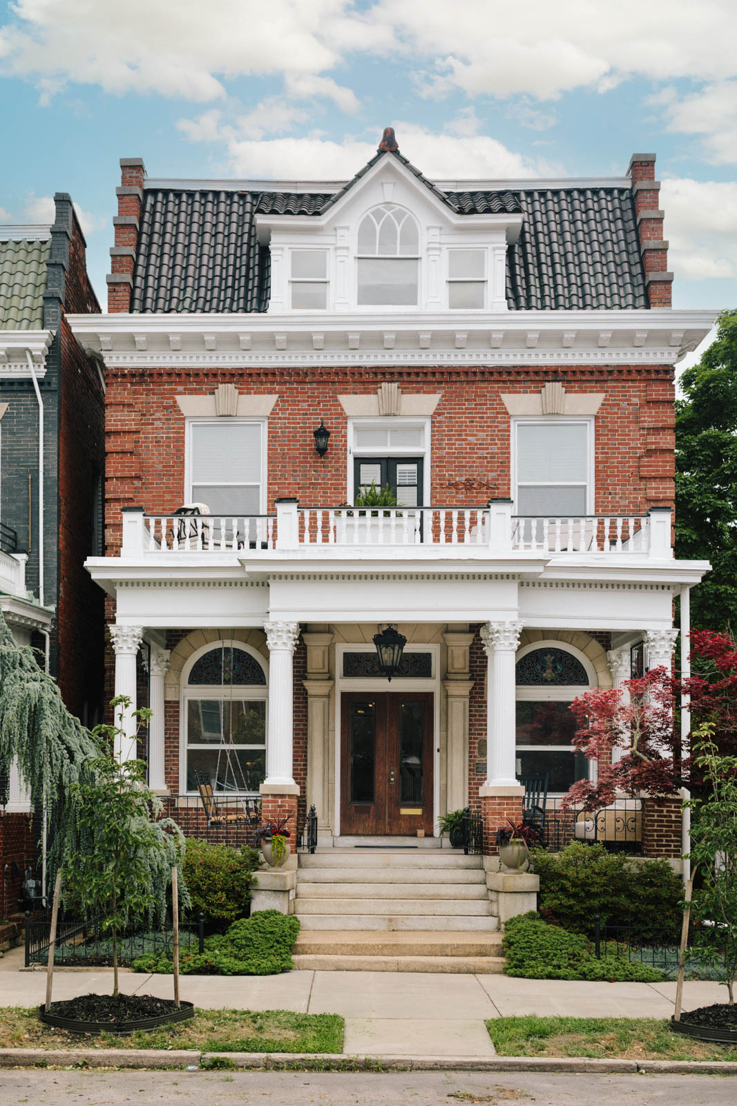 Three-story brick house with white decorative trim, columns, and a front porch, surrounded by trees and a sidewalk.