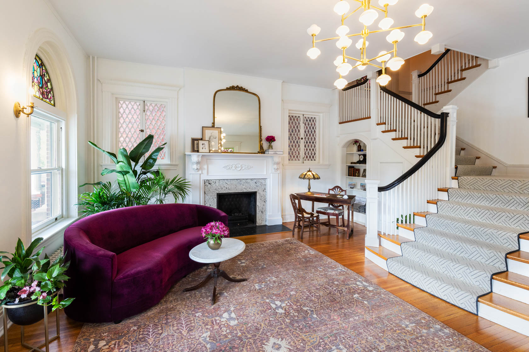 Living room with a purple velvet sofa, fireplace, staircase, and a small round table with flowers, bright natural light.