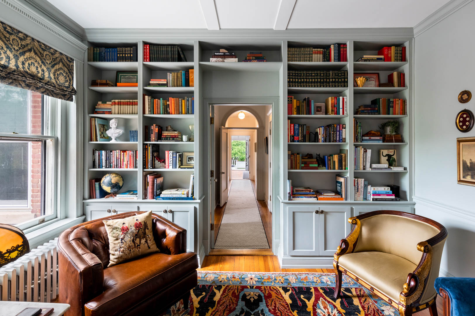 Living room with built-in bookshelves, a window with a patterned curtain, a leather armchair, and a patterned rug.